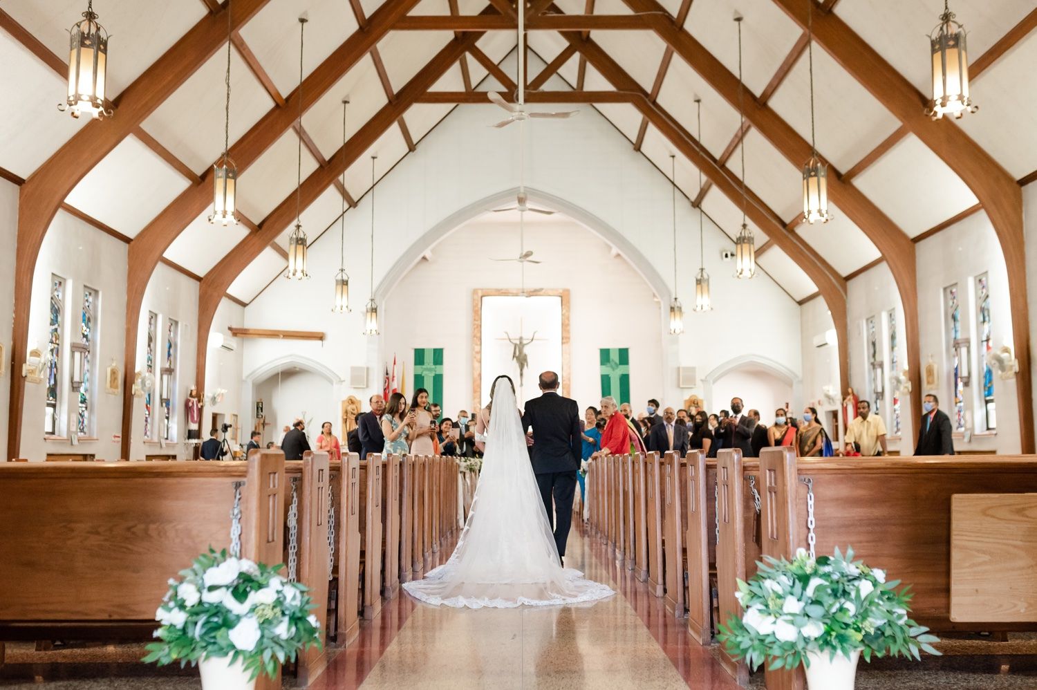 A traditional church wedding ceremony with wooden beam ceiling and guests seated in wooden pews adorned with white flowers.