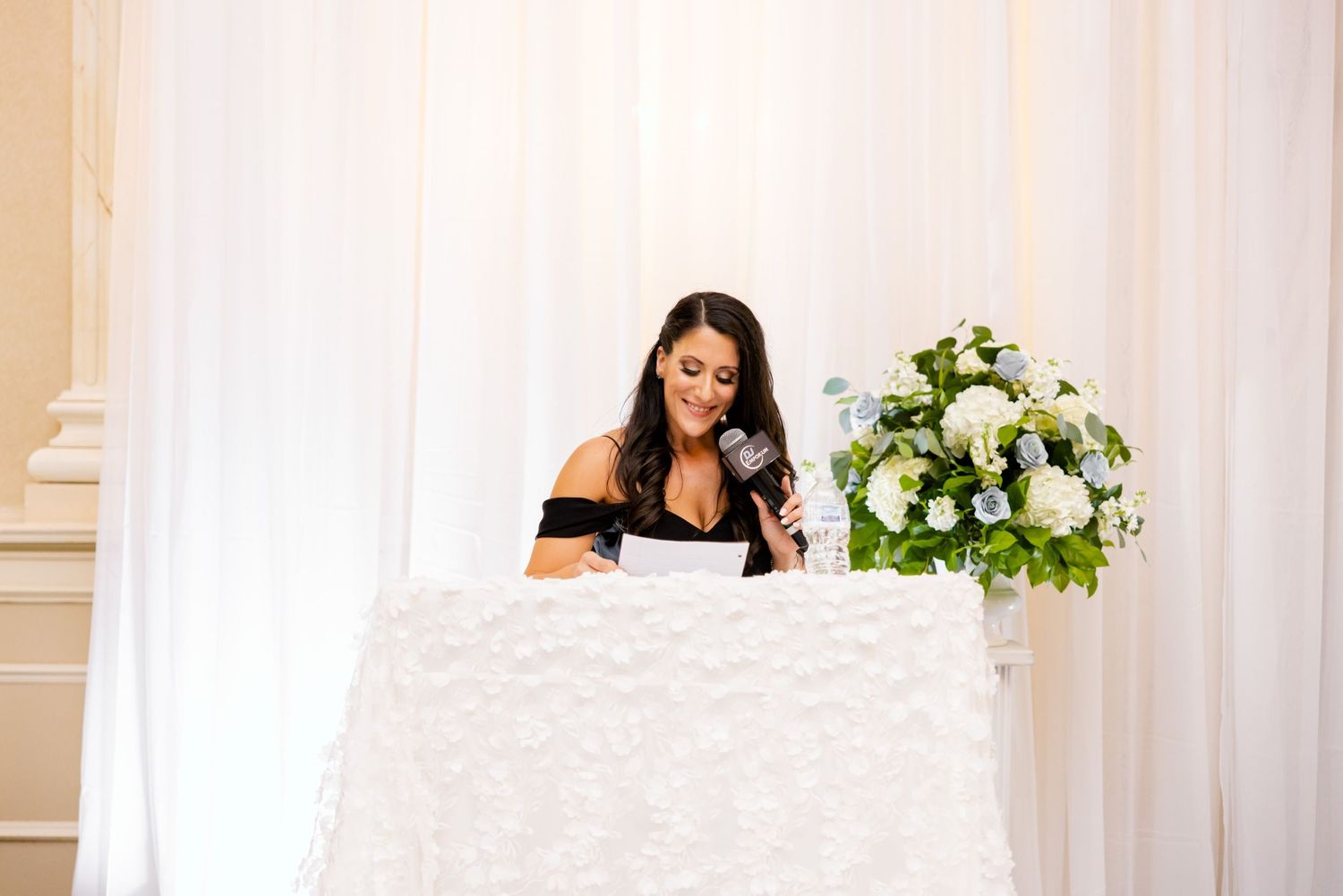 Speaker at wedding reception standing behind white linen covered table with floral arrangement.
