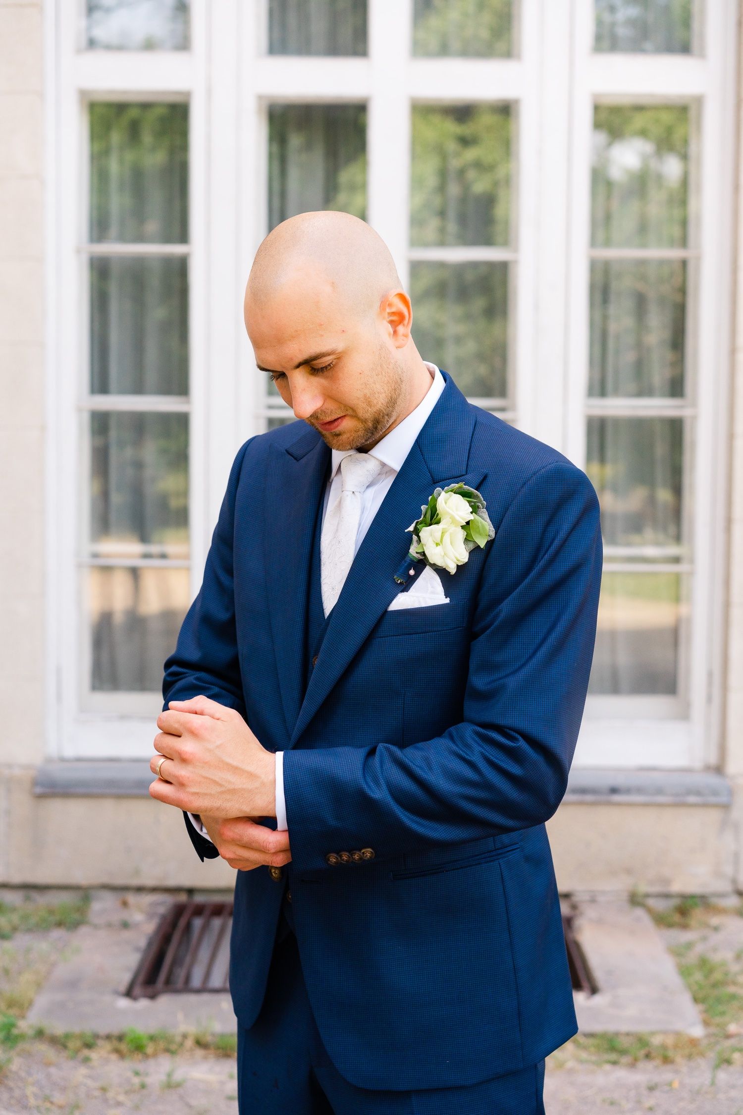 Groom adjusts sleeve of navy suit while standing against white building backdrop.