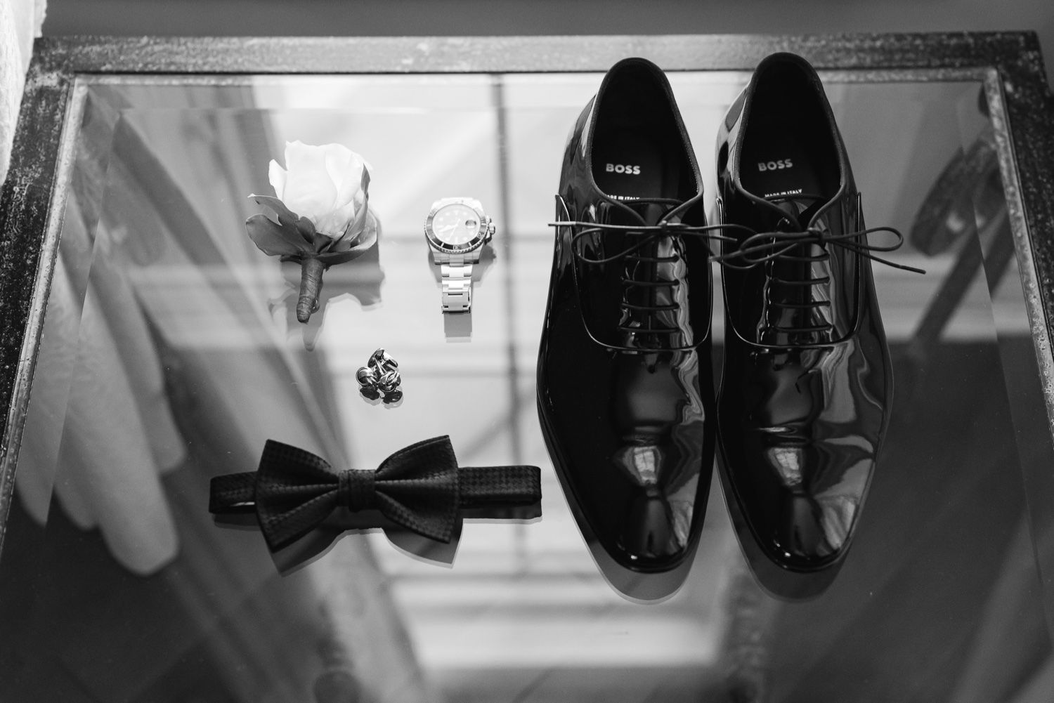 Wedding accessories including black dress shoes, bow tie, and watch displayed on glass surface.