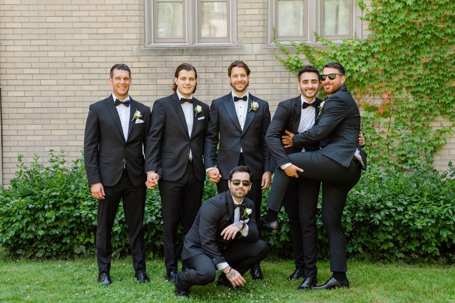 Groomsmen in black tuxedos pose for fun group photo with one member kneeling in front.