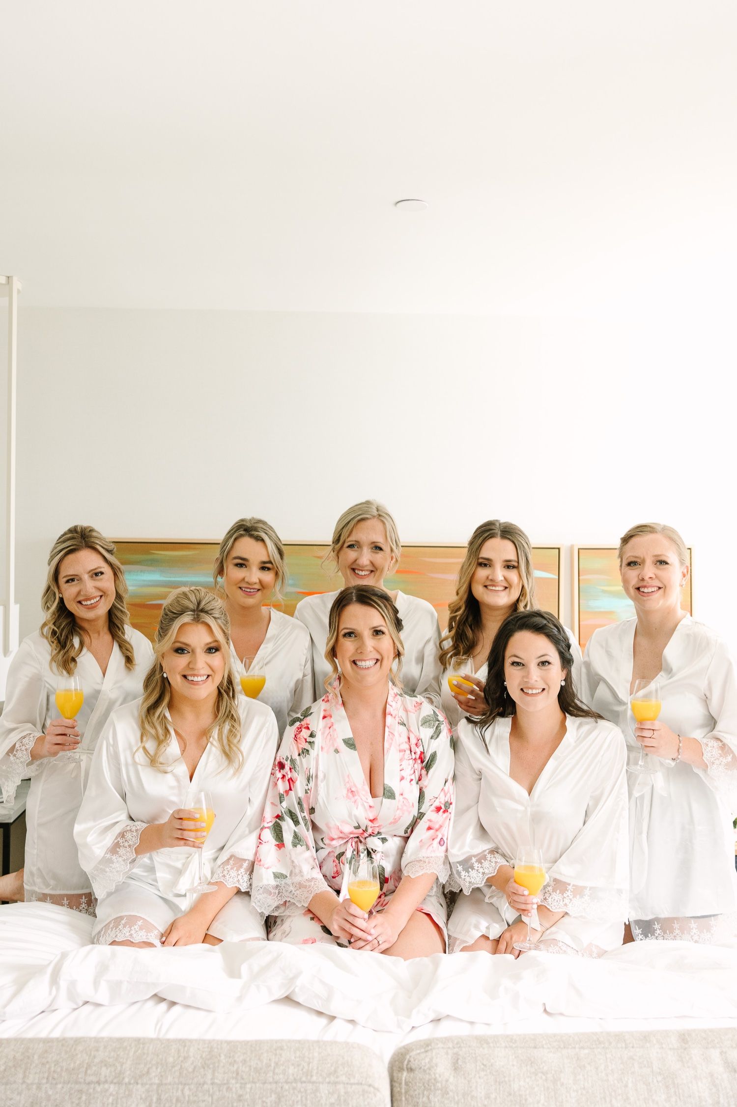 Bridesmaids in matching white robes sitting together on bed with mimosas.