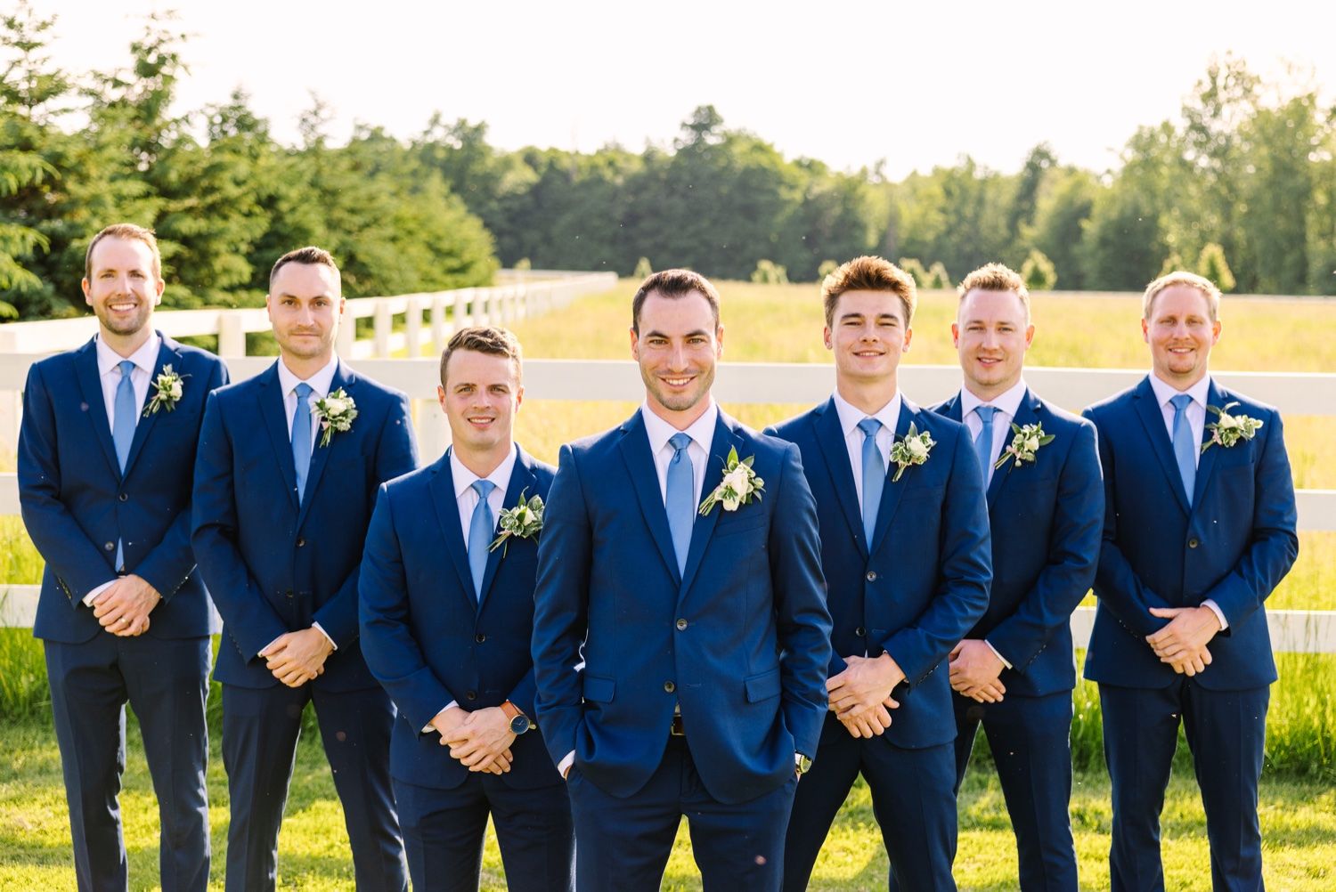 Groomsmen in coordinated navy suits pose together in sunlit outdoor setting.