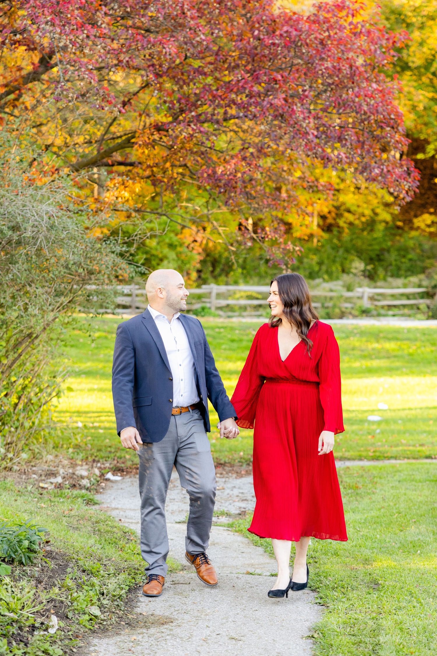 Two people stroll hand in hand on a path beneath colorful fall trees with red and yellow leaves.