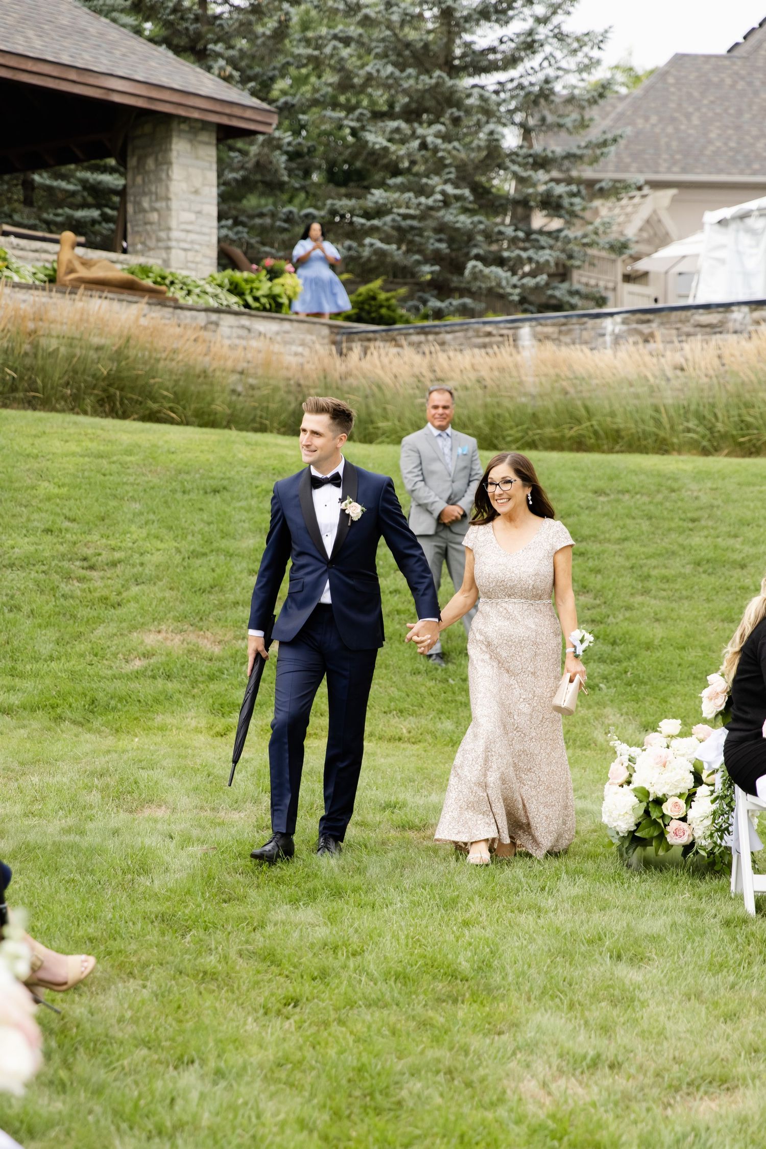 Wedding party walks across grassy lawn with elegant house in background.