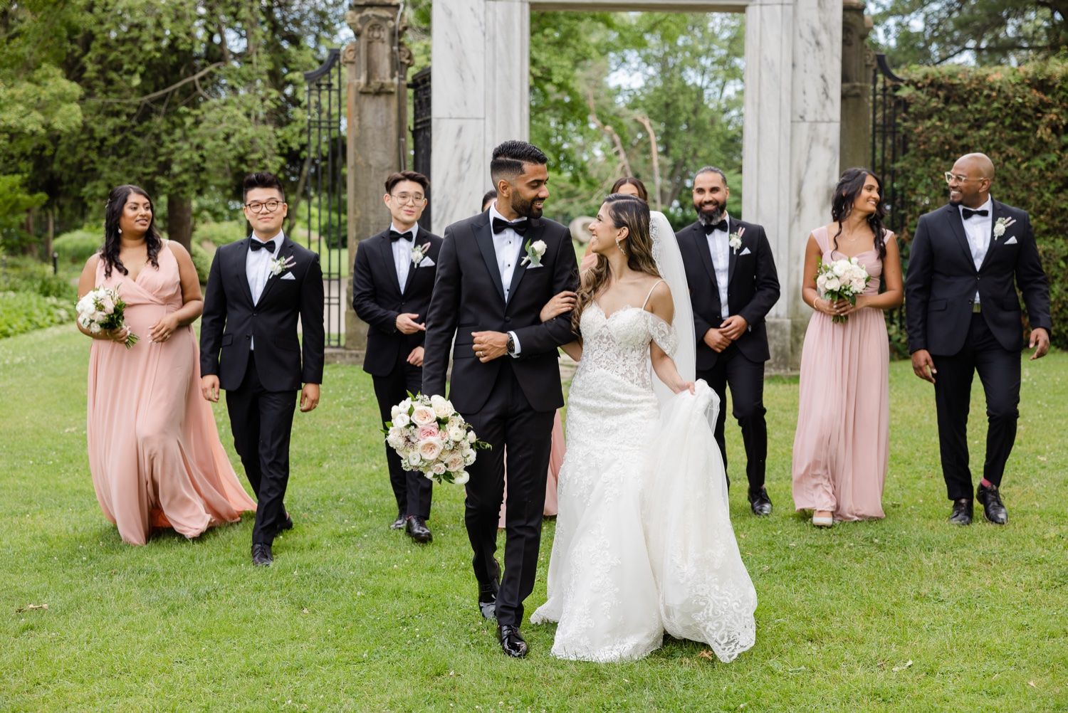 Wedding party walking together on grass in front of a classical stone archway.
