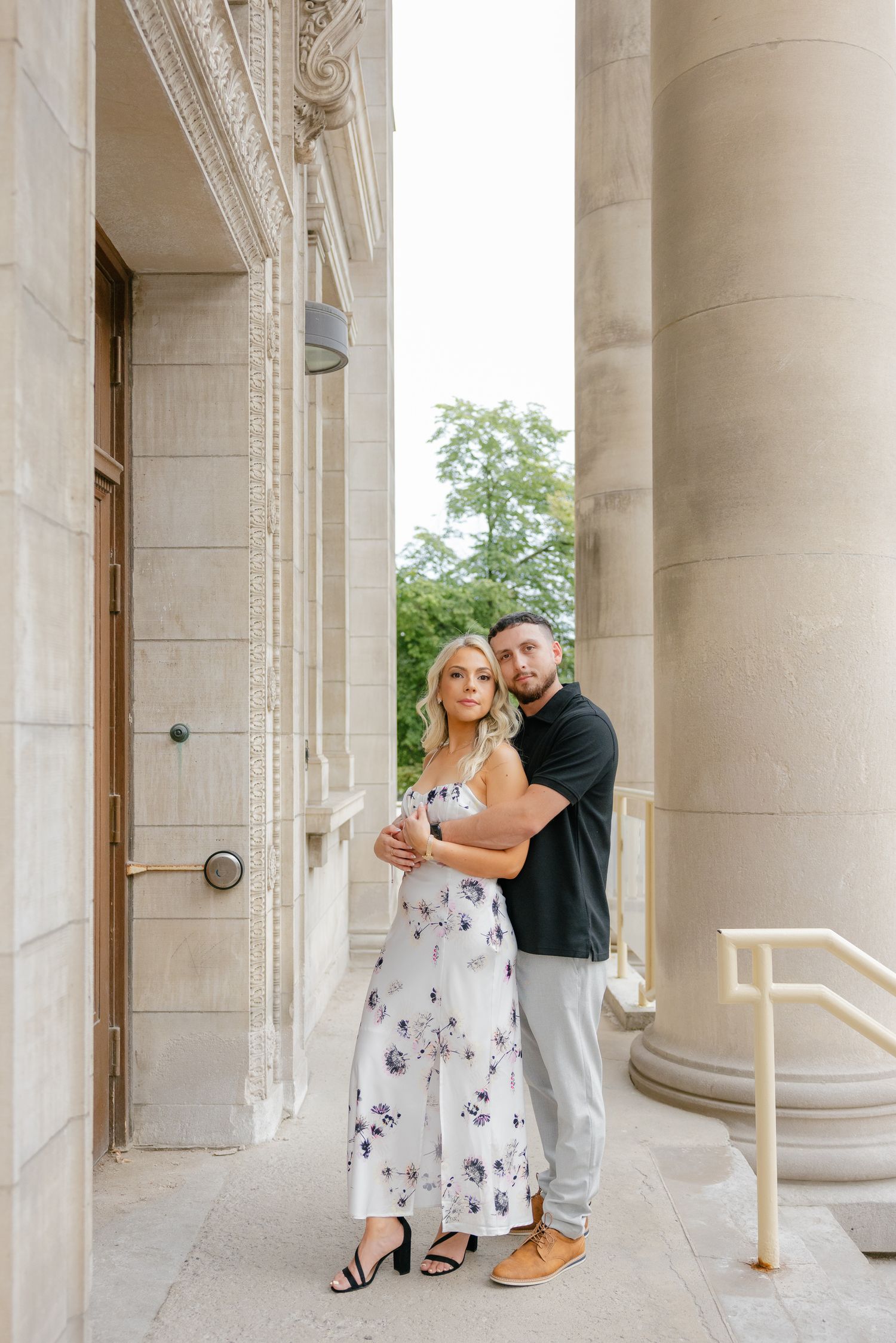 A romantic couple poses between elegant stone columns, with her wearing a floral dress and him in a black polo.