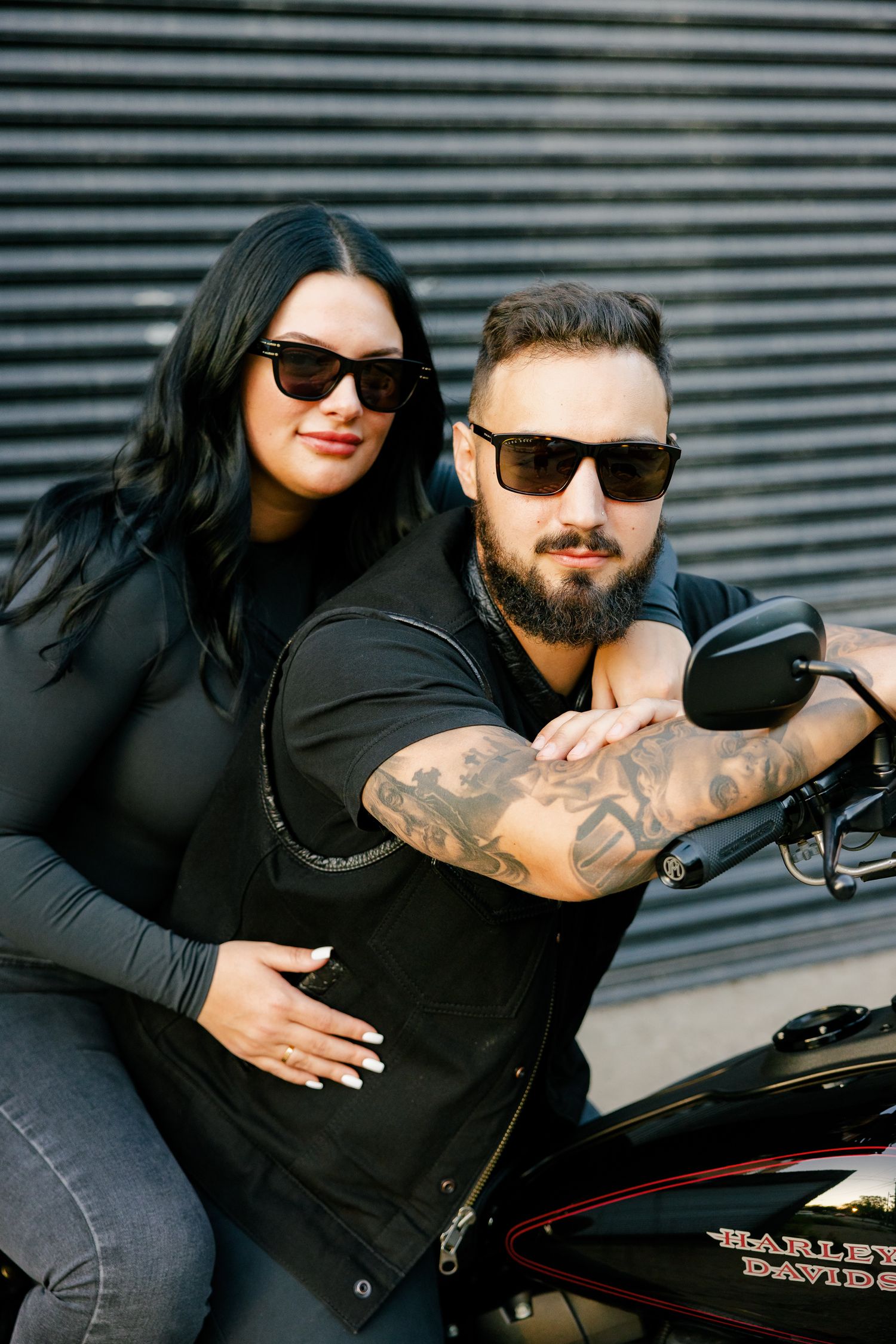 Couple in sunglasses poses together on motorcycle against industrial background.