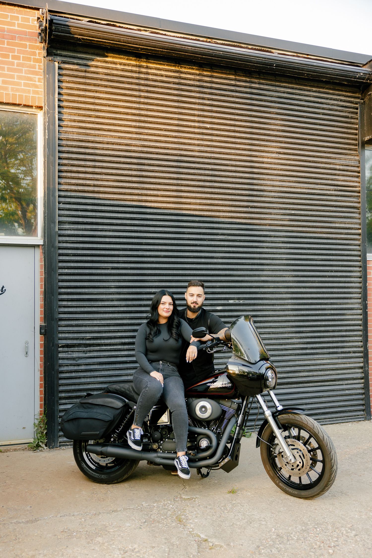 A couple sits on their black motorcycle against a metal roll-up door.