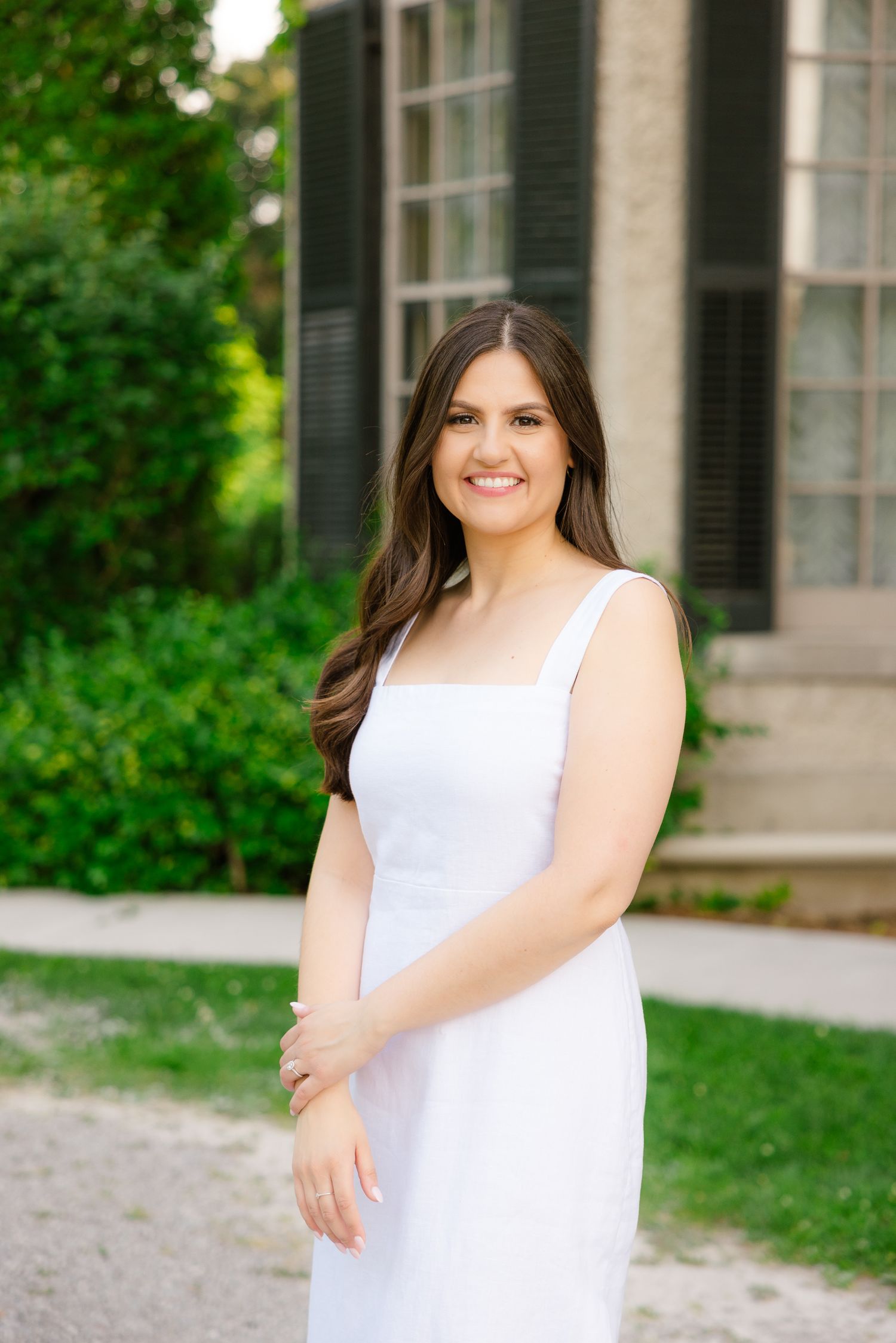 Woman in elegant white dress poses by historic mansion with black shutters.