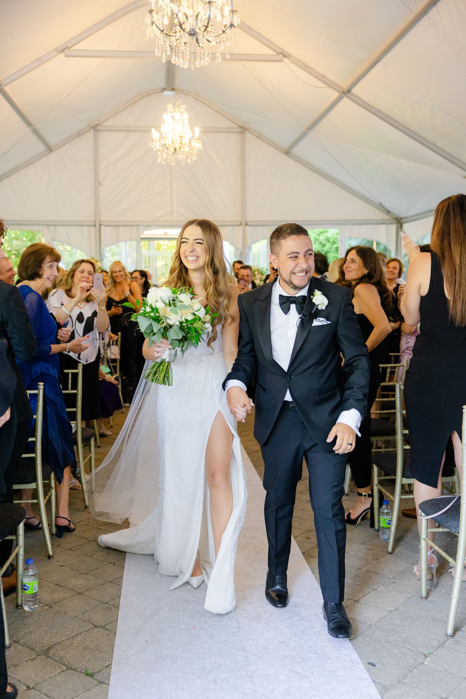 Celebratory recessional walk of newlyweds down a decorated tent aisle with guests and chandelier overhead.