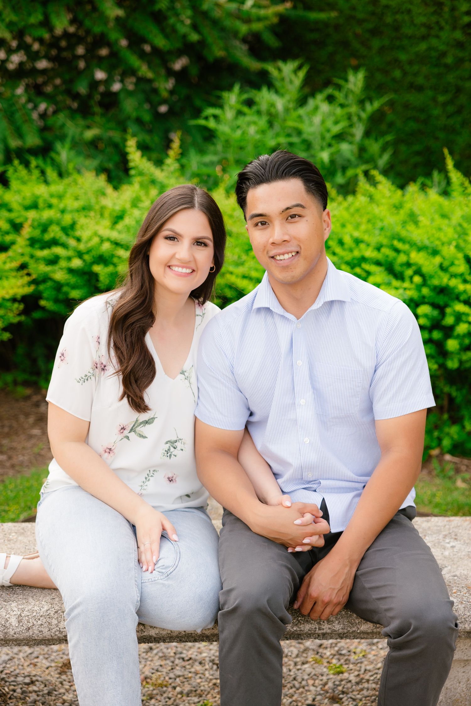 A couple smiles at the camera while sitting on a stone bench in front of green landscaping.