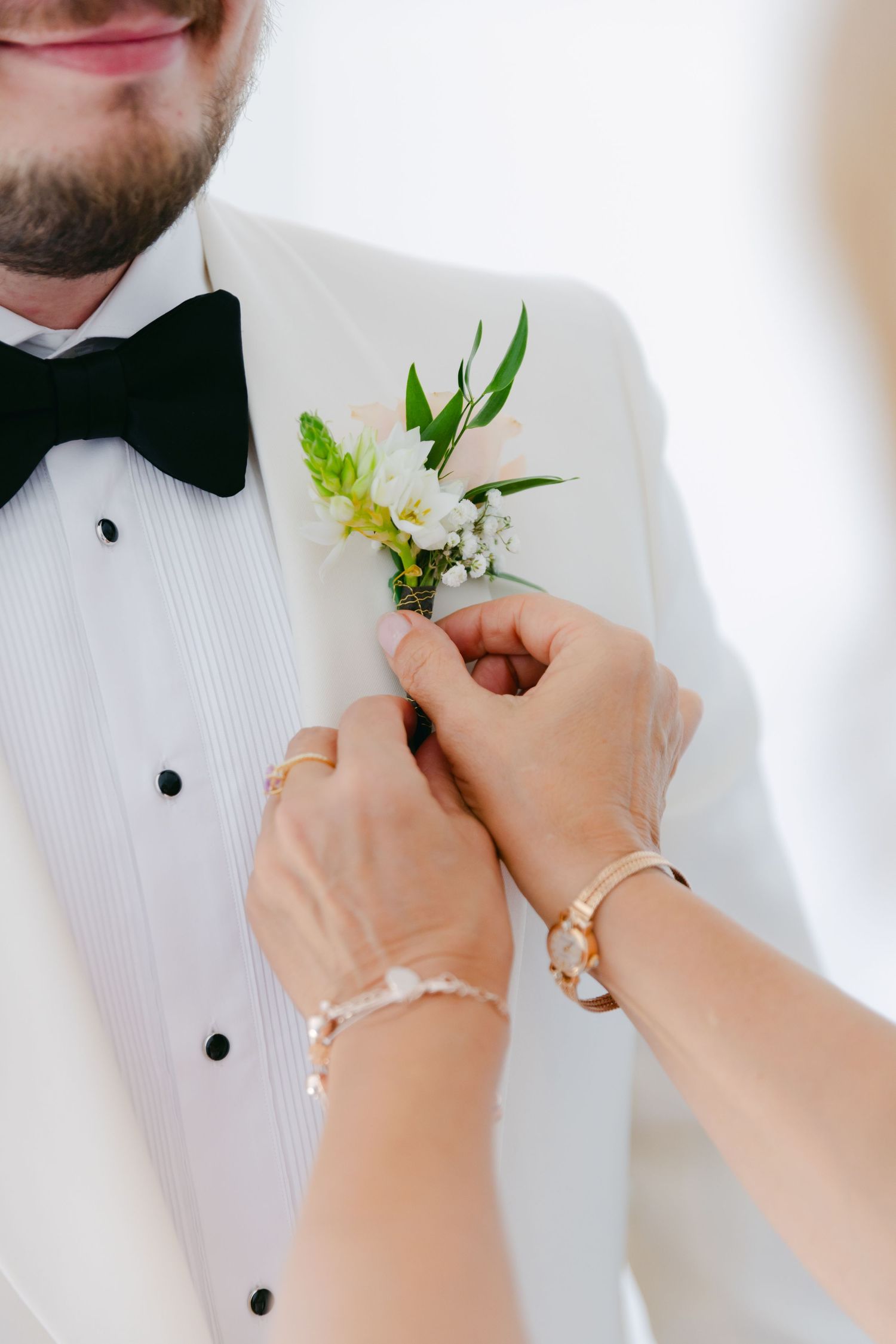 Hands pinning a delicate white floral boutonniere onto a formal tuxedo lapel.