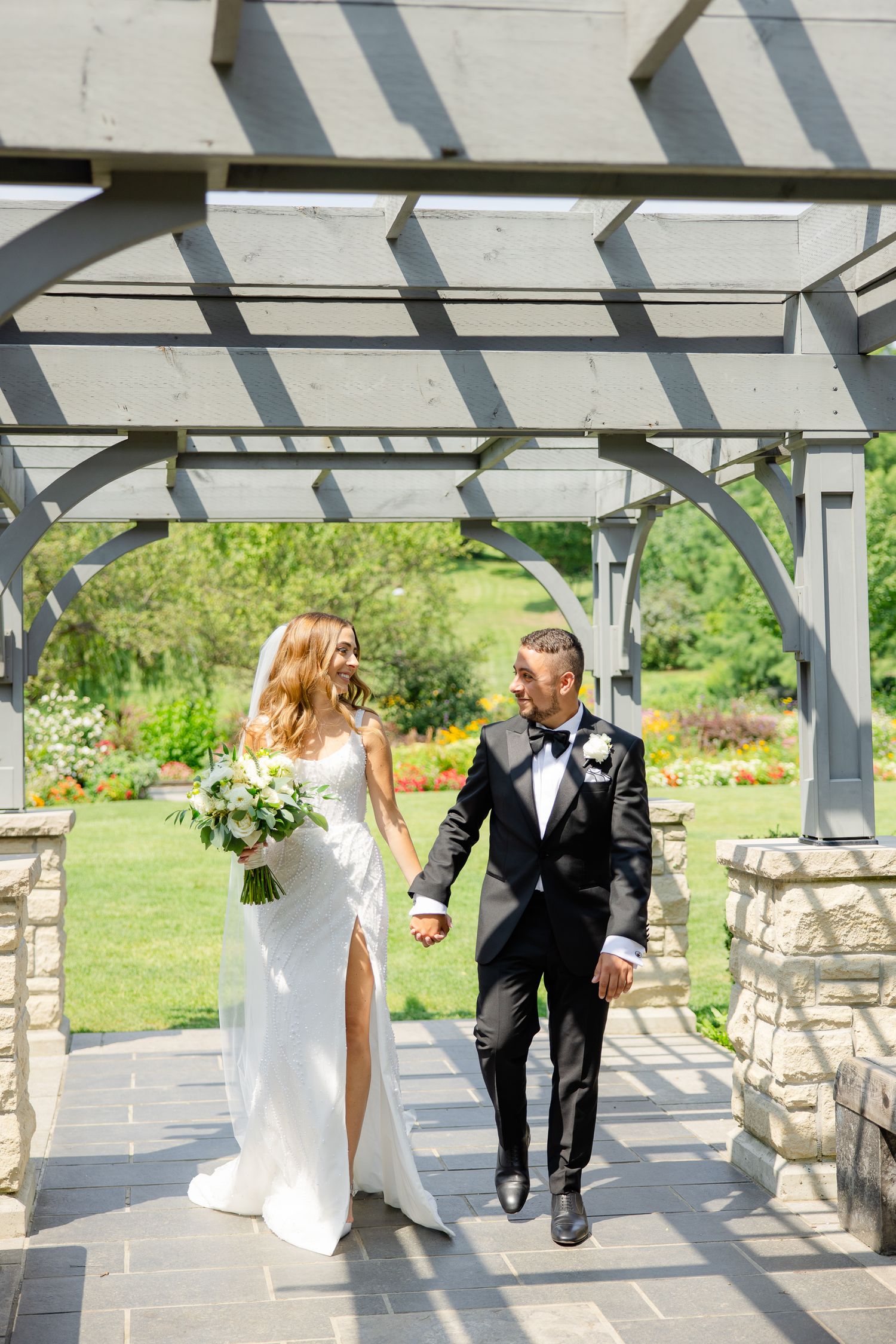 Newlyweds walk hand in hand under a pergola with beautiful garden backdrop.