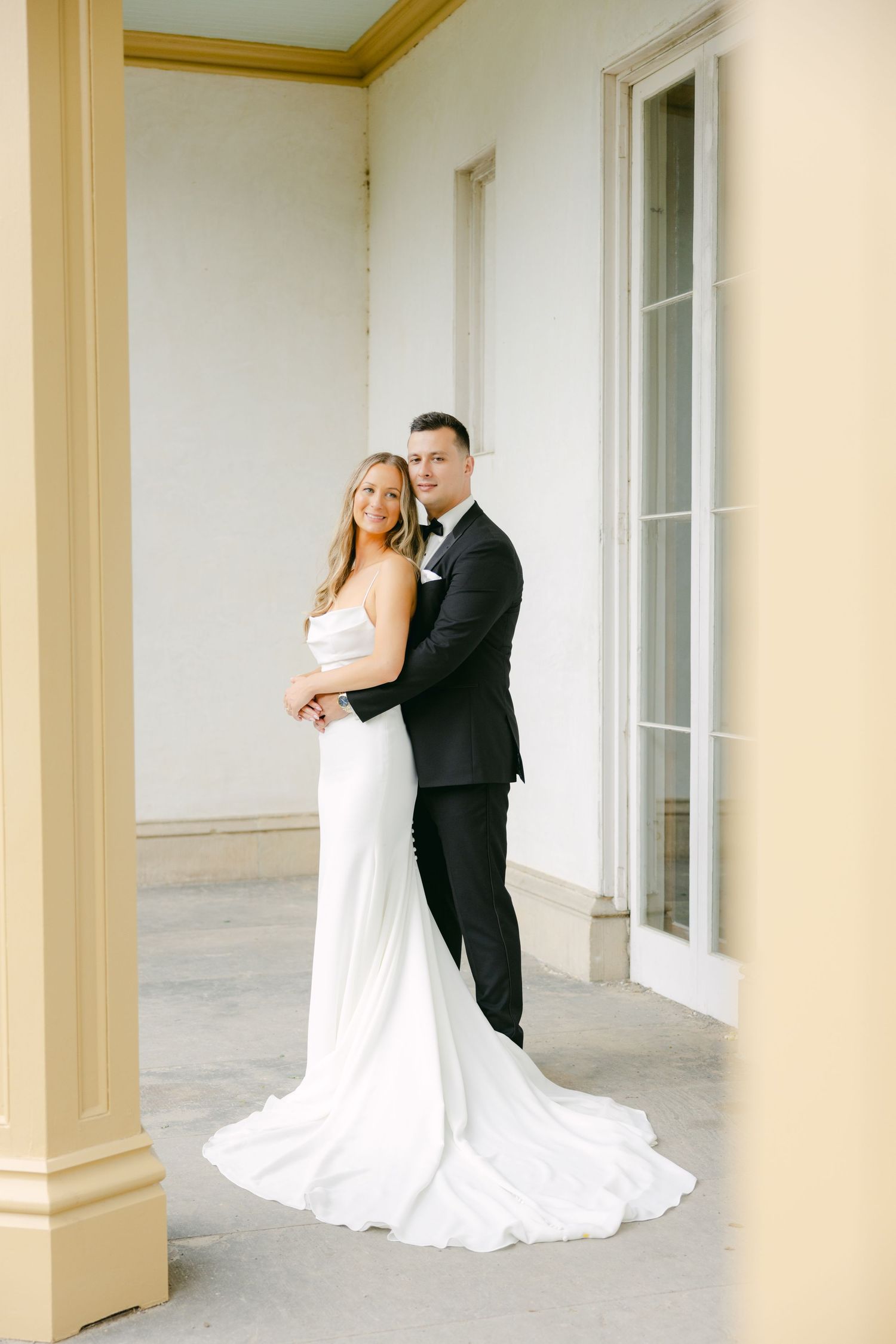 Newlyweds in formal attire standing together on classic columned porch.