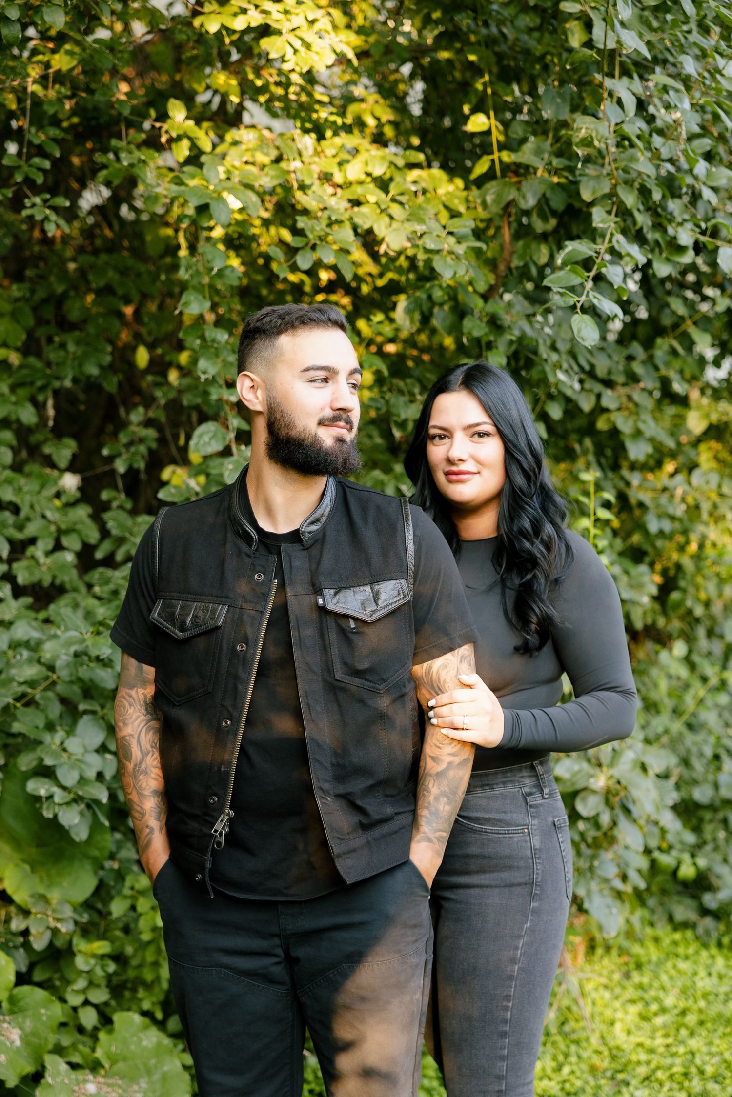 A stylish couple poses together in a park with dappled sunlight filtering through the trees.
