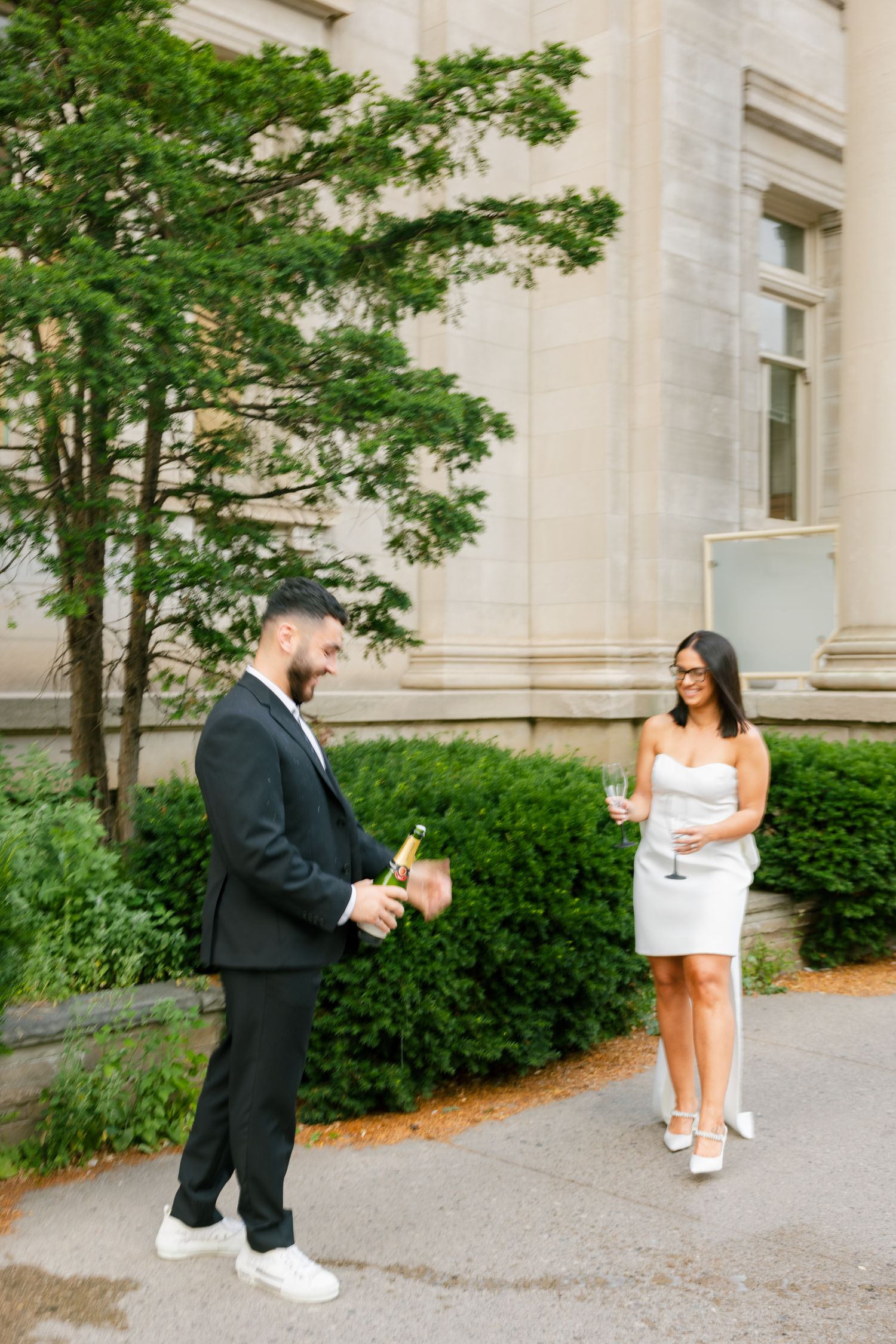 A couple shares a celebratory moment with drinks outside a grand building with landscaping.