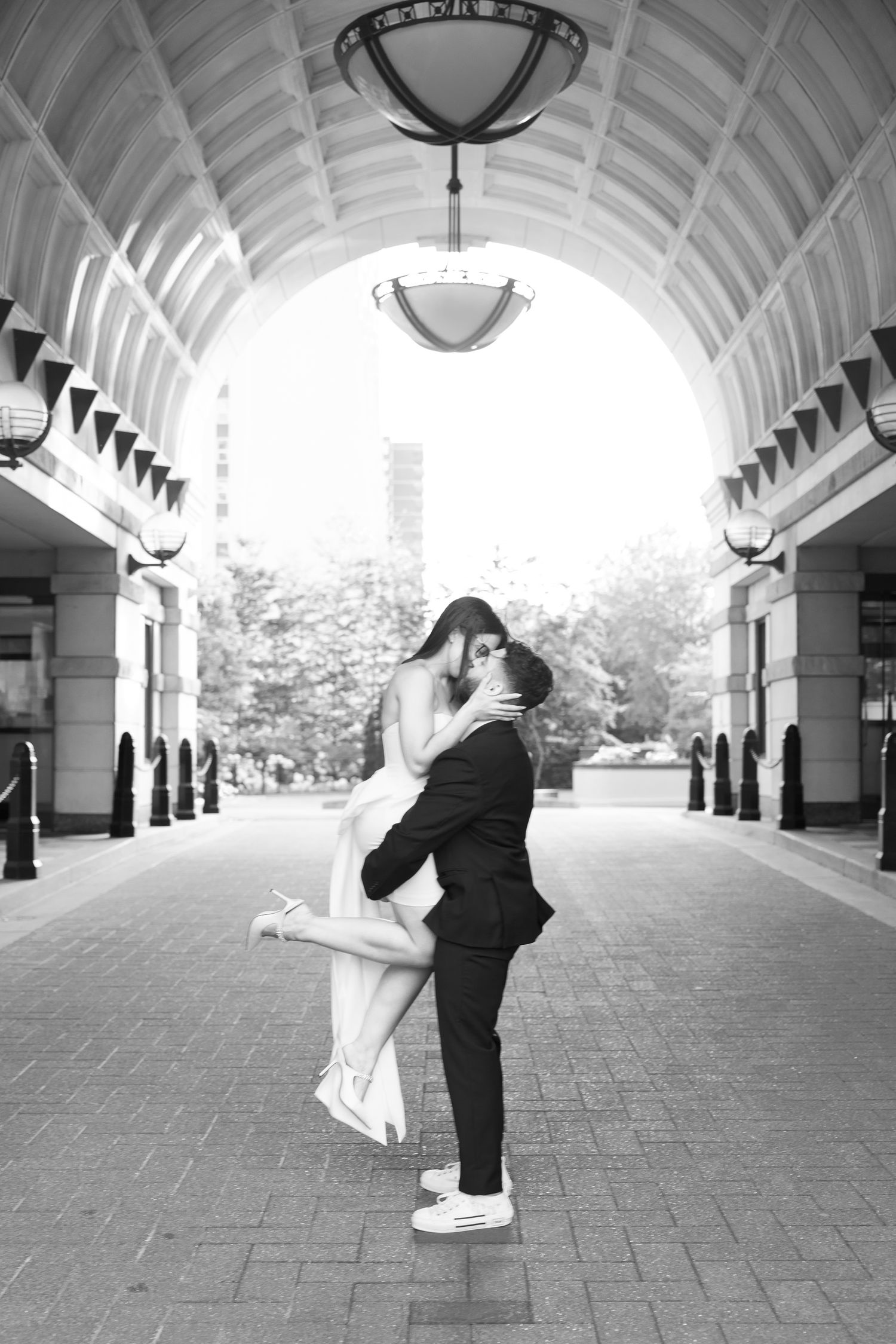 Black and white photograph of an intimate embrace under a vaulted ceiling with pendant lights.