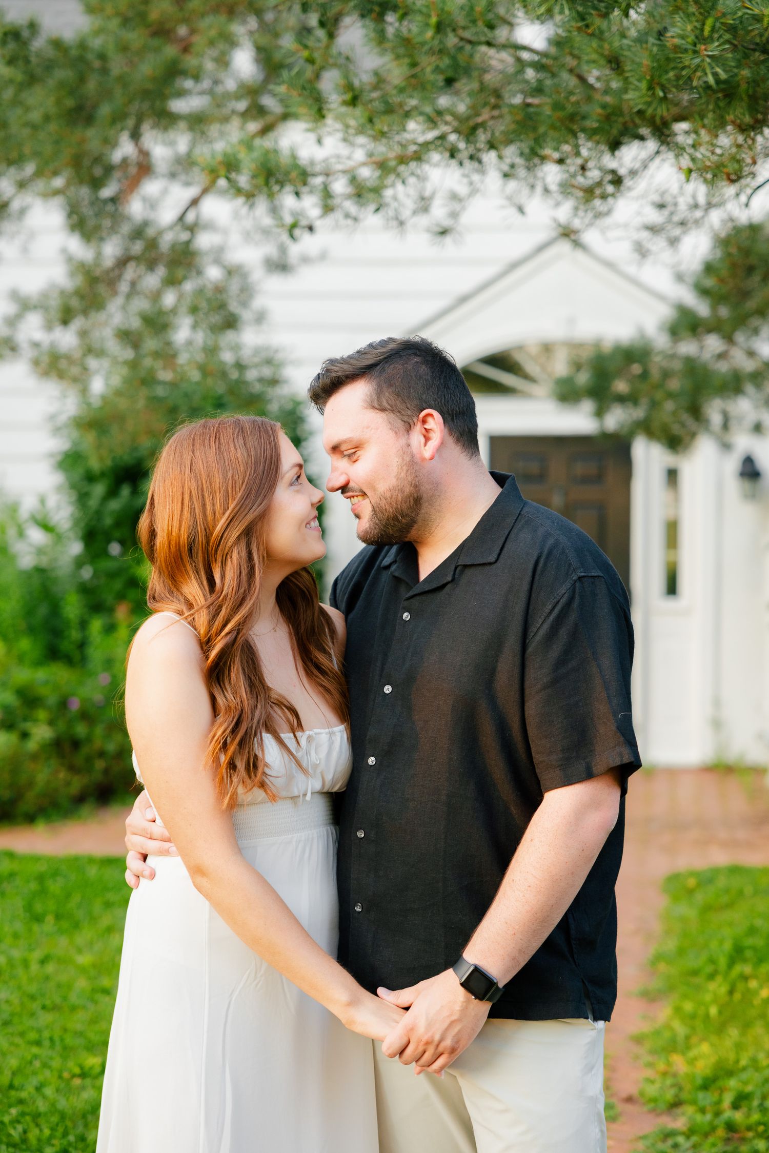 Two people embrace while standing on a brick path with a classic white house in the background.