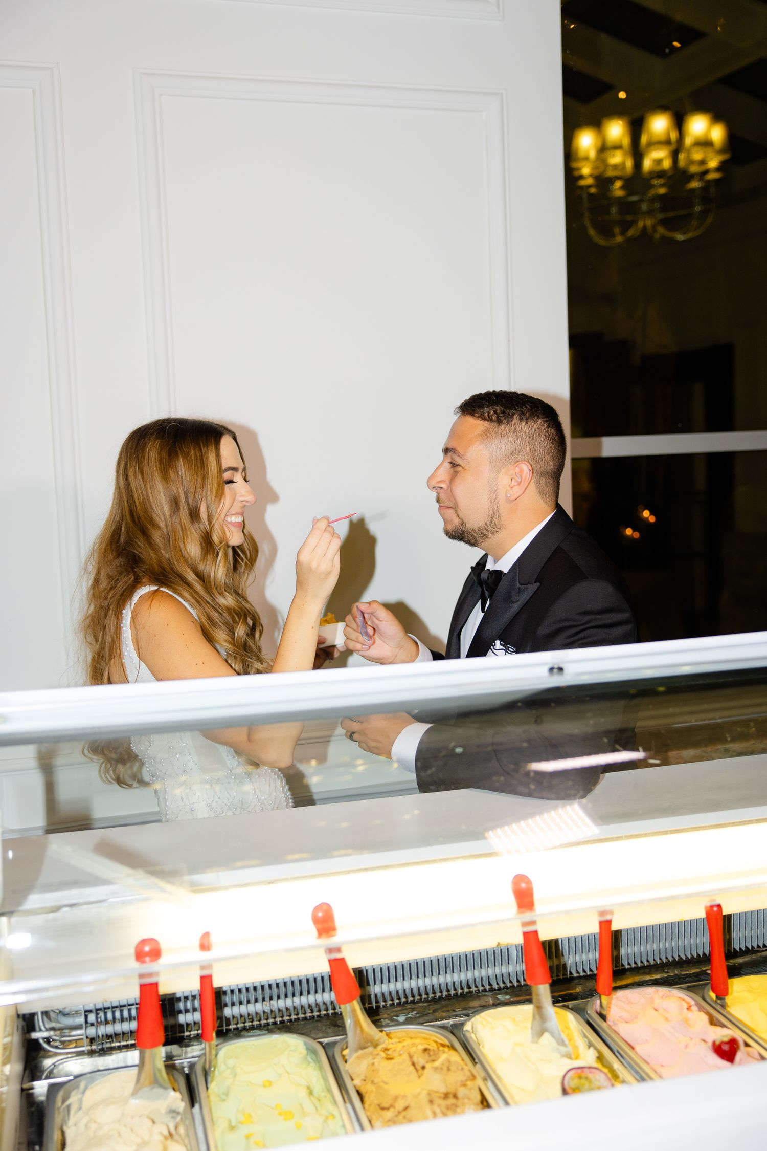 Couple laughing while sharing gelato from an elegant display cart at formal event.