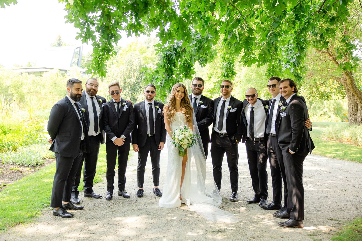 Wedding party in black suits posing with couple on garden path surrounded by green trees.