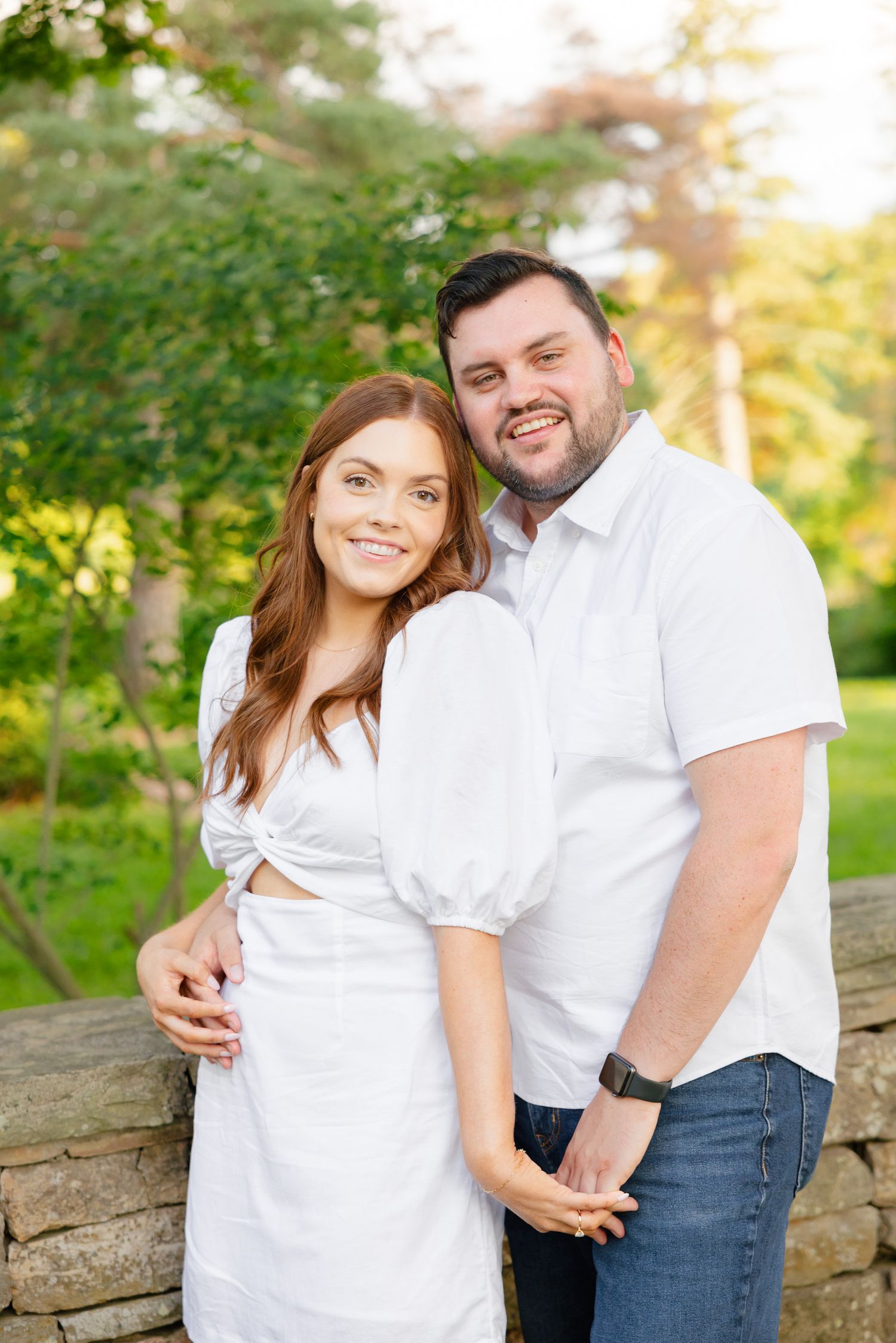 A couple in coordinating white outfits smiles at the camera while posing by a garden wall.