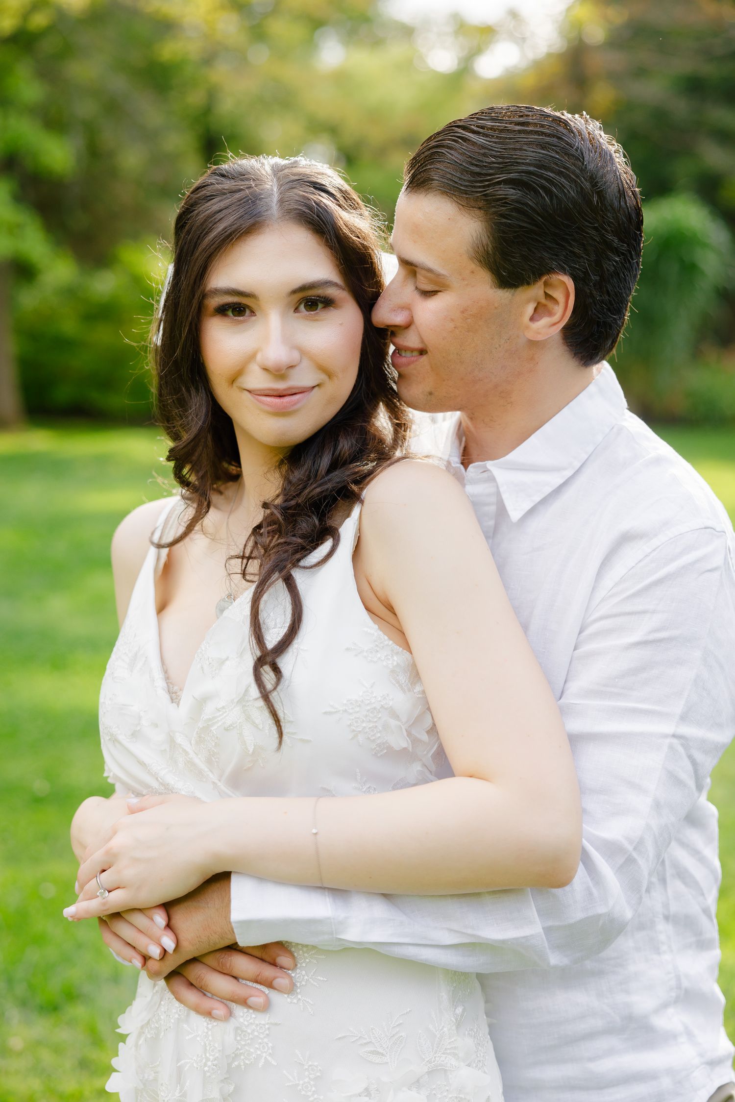 Romantic outdoor engagement portrait of couple embracing in white outfits against green garden background.