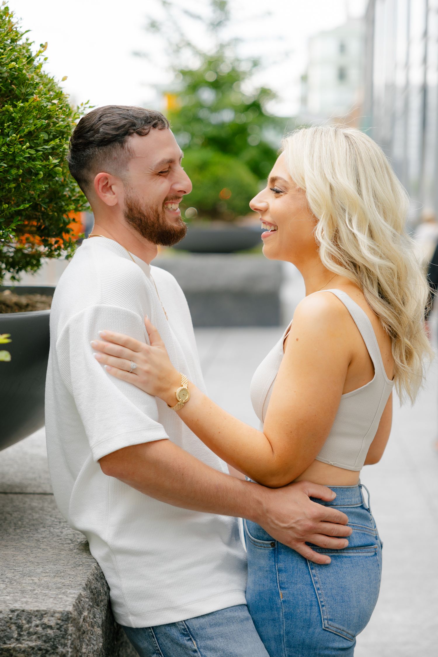 A laughing couple embraces near greenery and modern architecture.