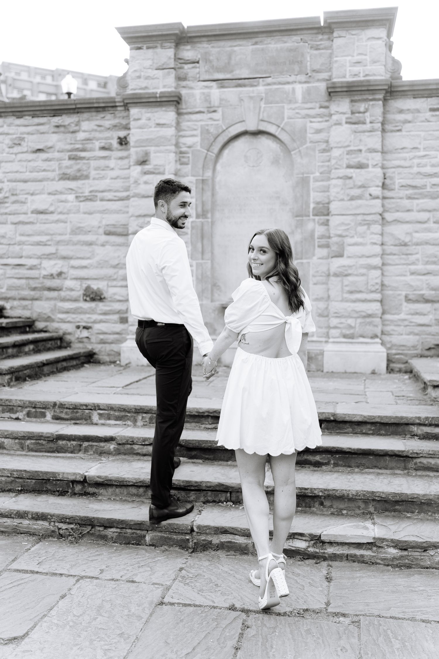 A black and white engagement photo of a couple posing on historic stone steps with architectural details.