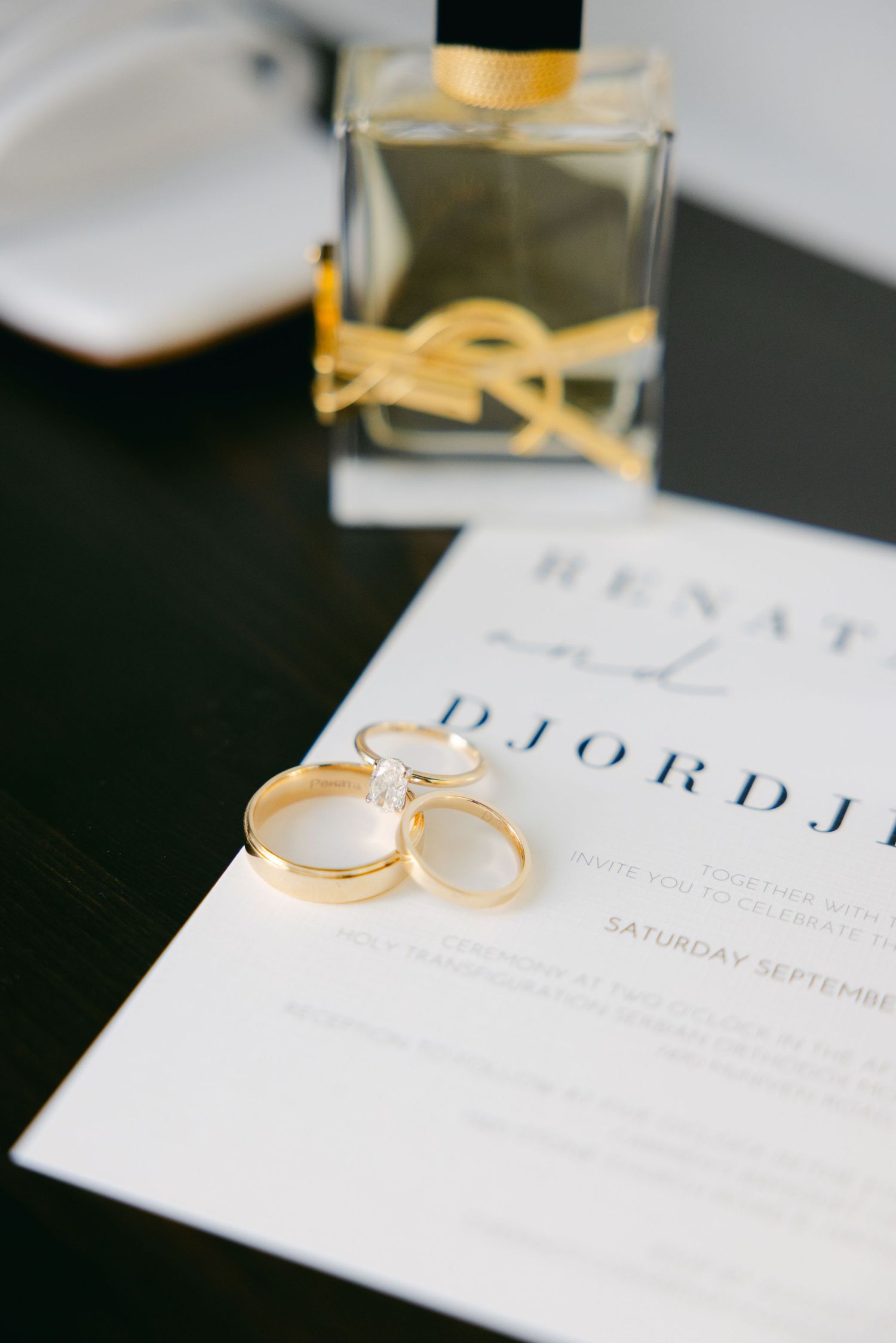 Wedding rings and perfume bottle arranged on an elegant wedding invitation.