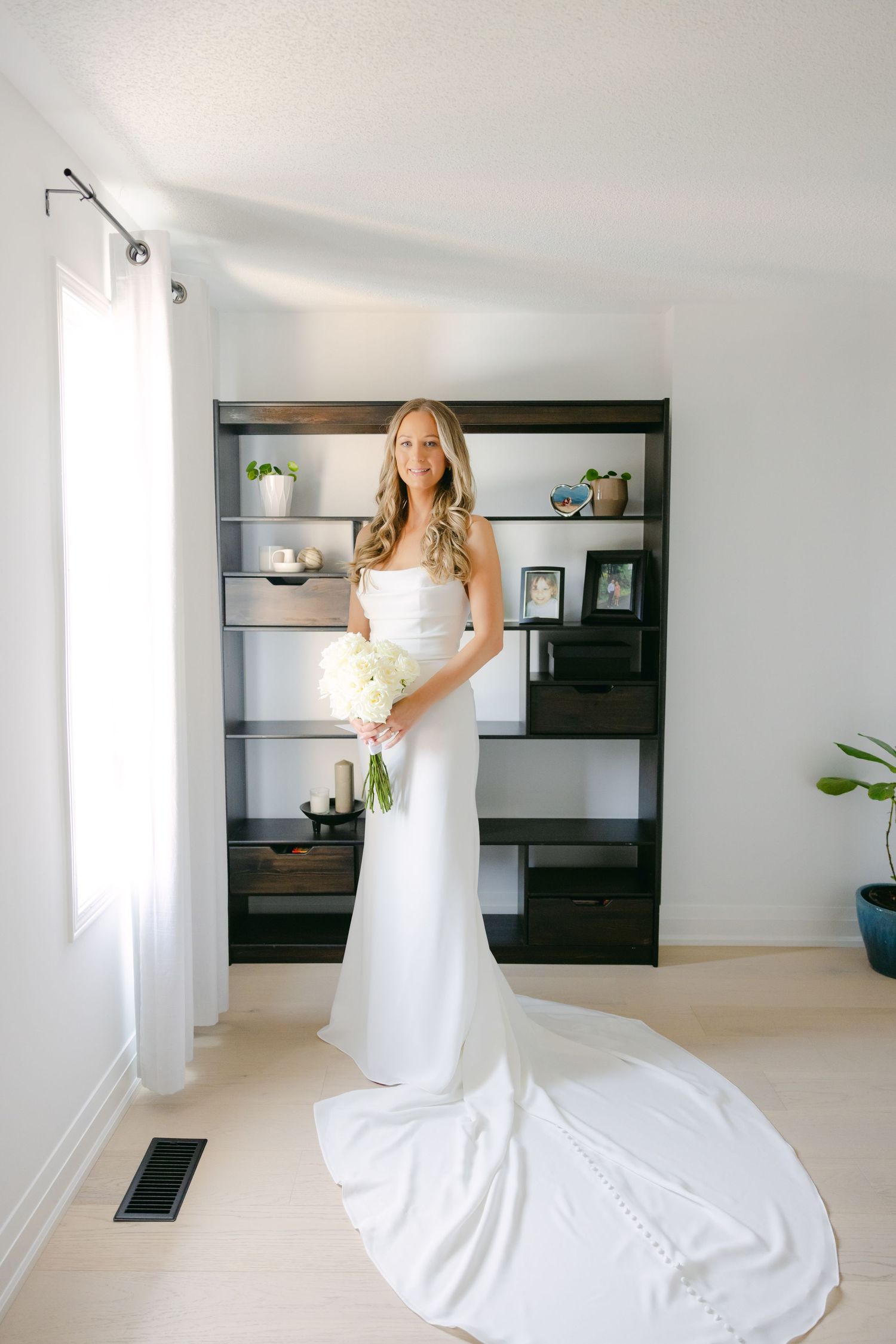 Elegant white wedding dress with long train displayed in modern room with built-in shelving.