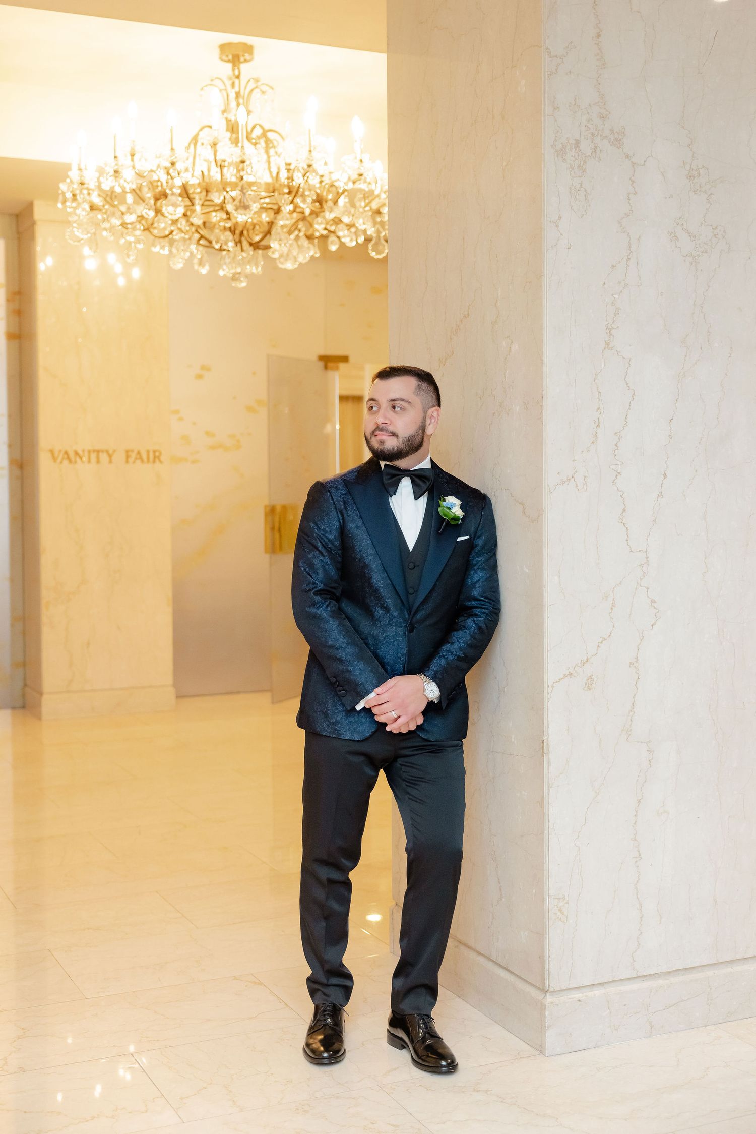 Groomsmen in dark suits stand in a marble-walled venue with crystal chandeliers.