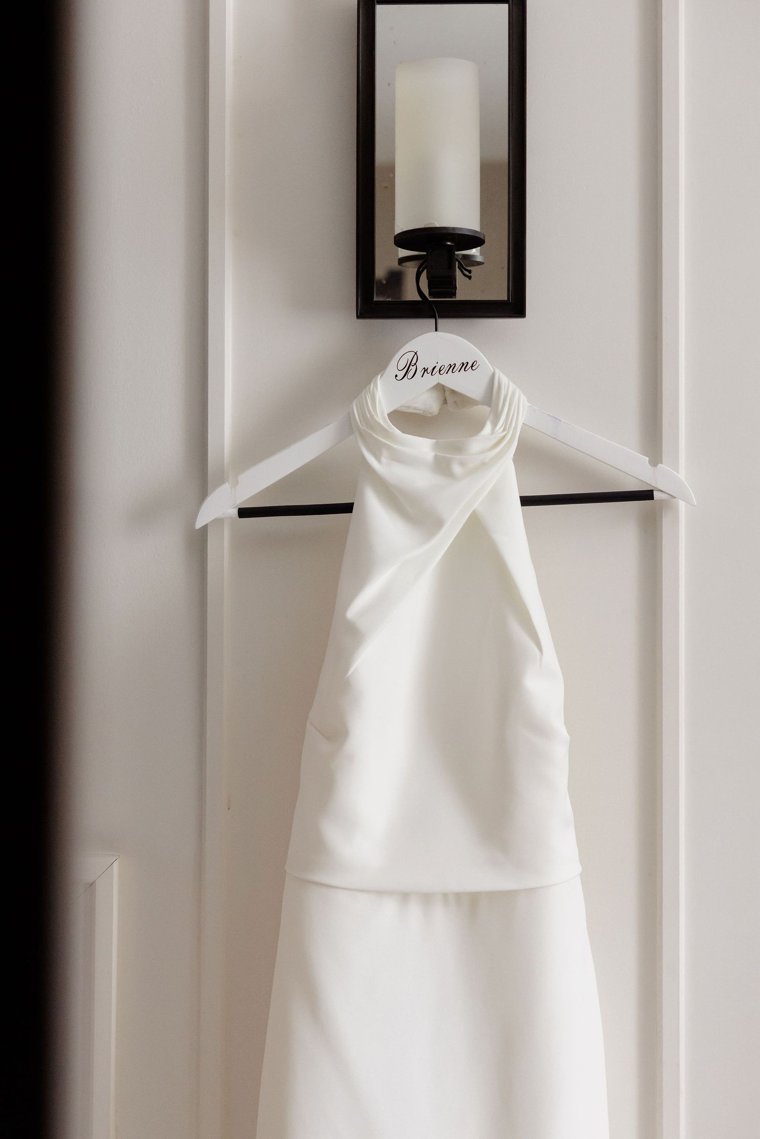 A white halter-style wedding dress hanging on display against a white wall with black frames.