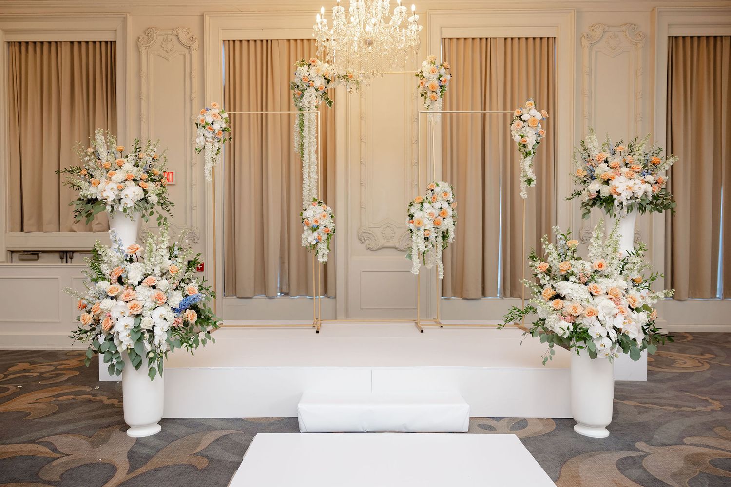 Elegant wedding ceremony backdrop with white floral arrangements and crystal chandelier in a luxurious ballroom setting.