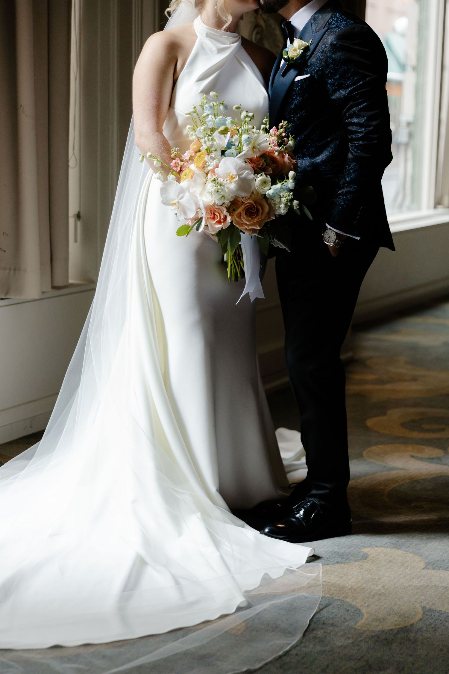 Elegant wedding couple embraces in classic white gown and dark suit against neutral backdrop.