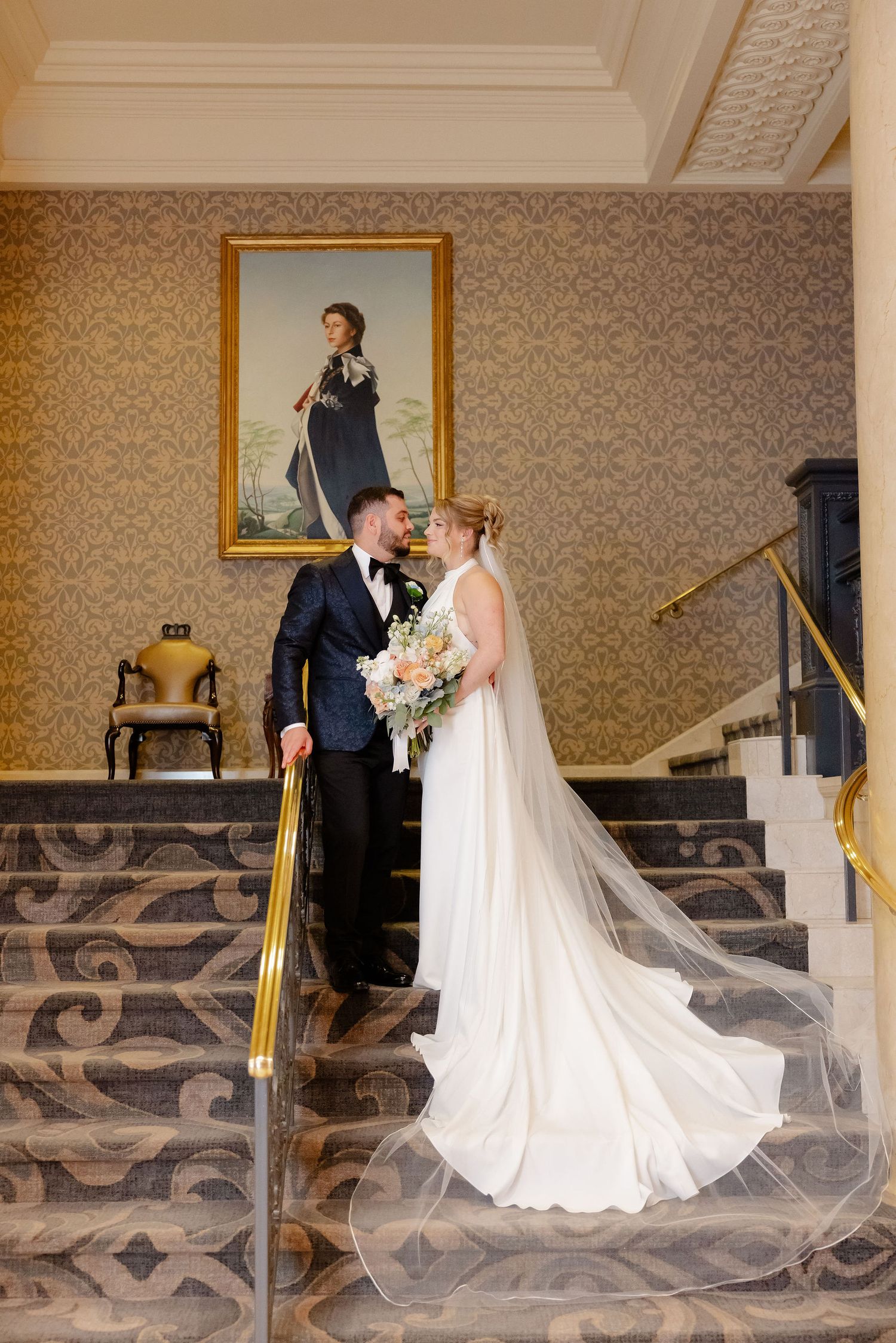 Wedding couple poses on ornate staircase with decorative wallpaper and vintage portrait.