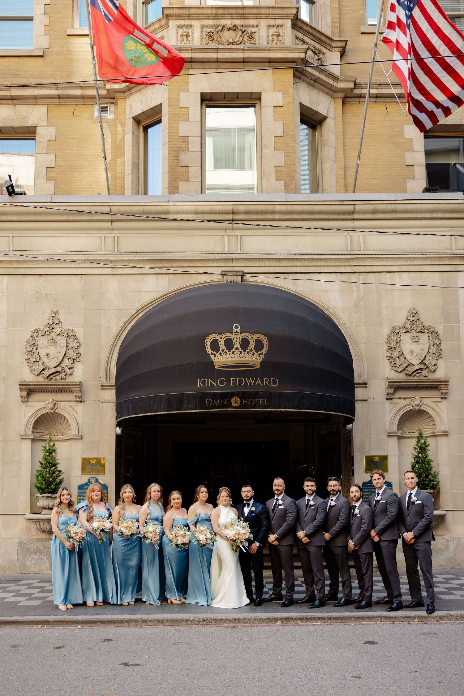 Wedding party in blue dresses and dark suits poses outside the King Edward Hotel entrance.