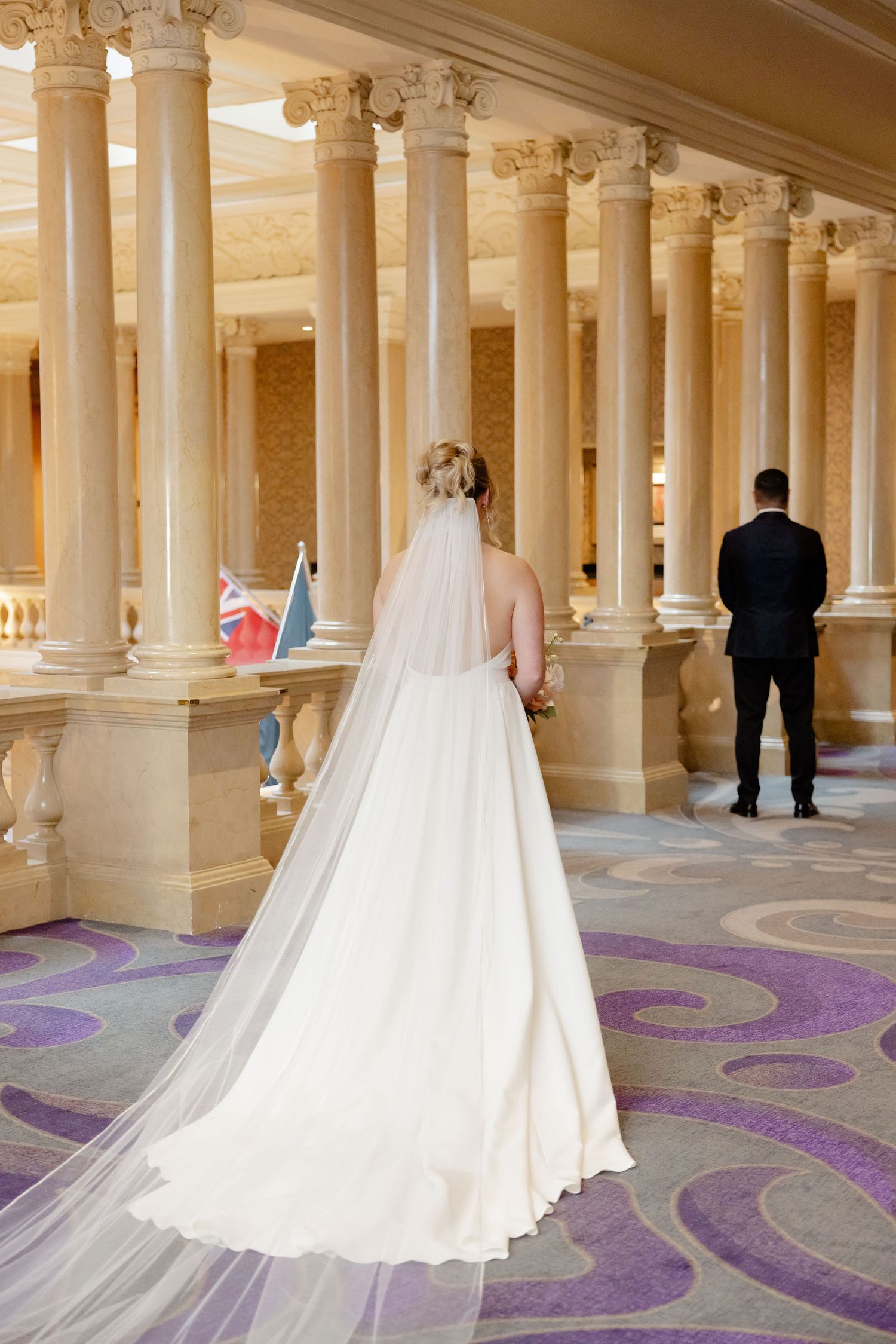 A wedding dress train flows elegantly across a grand colonnade with purple carpet patterns.