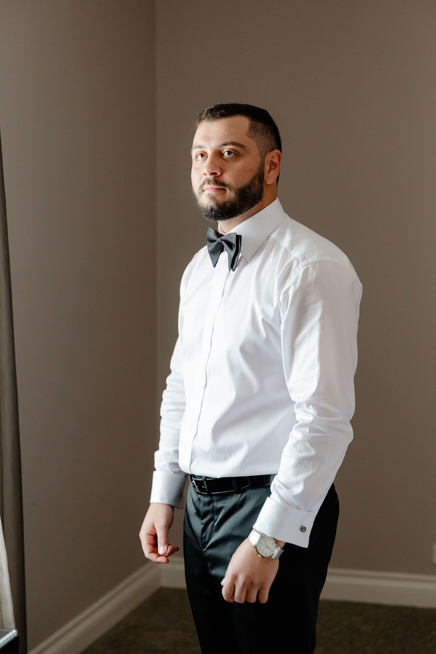 A groom in a crisp white dress shirt and black bow tie stands against a neutral wall.