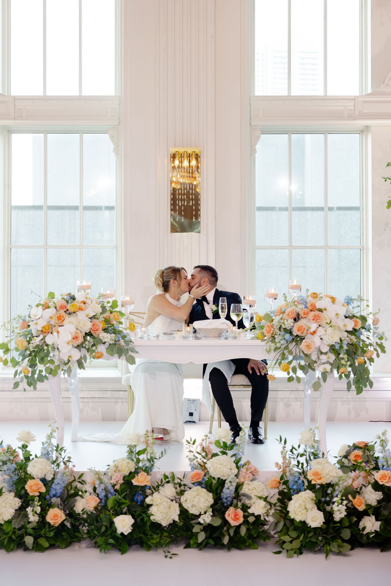 Wedding couple seated at decorated head table with lush floral arrangements.