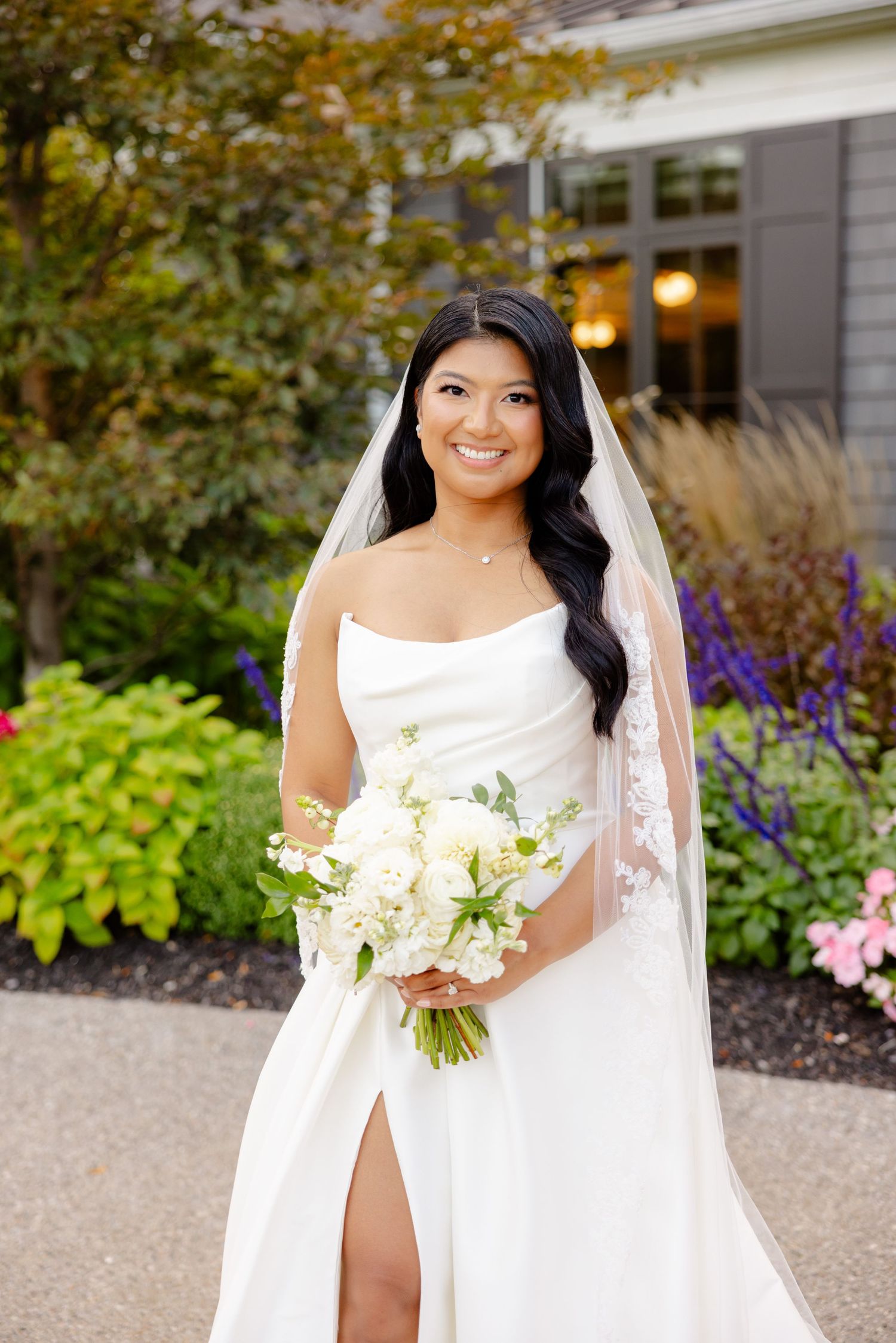 White wedding bouquet complements sleek bridal gown in garden setting with purple flowers.