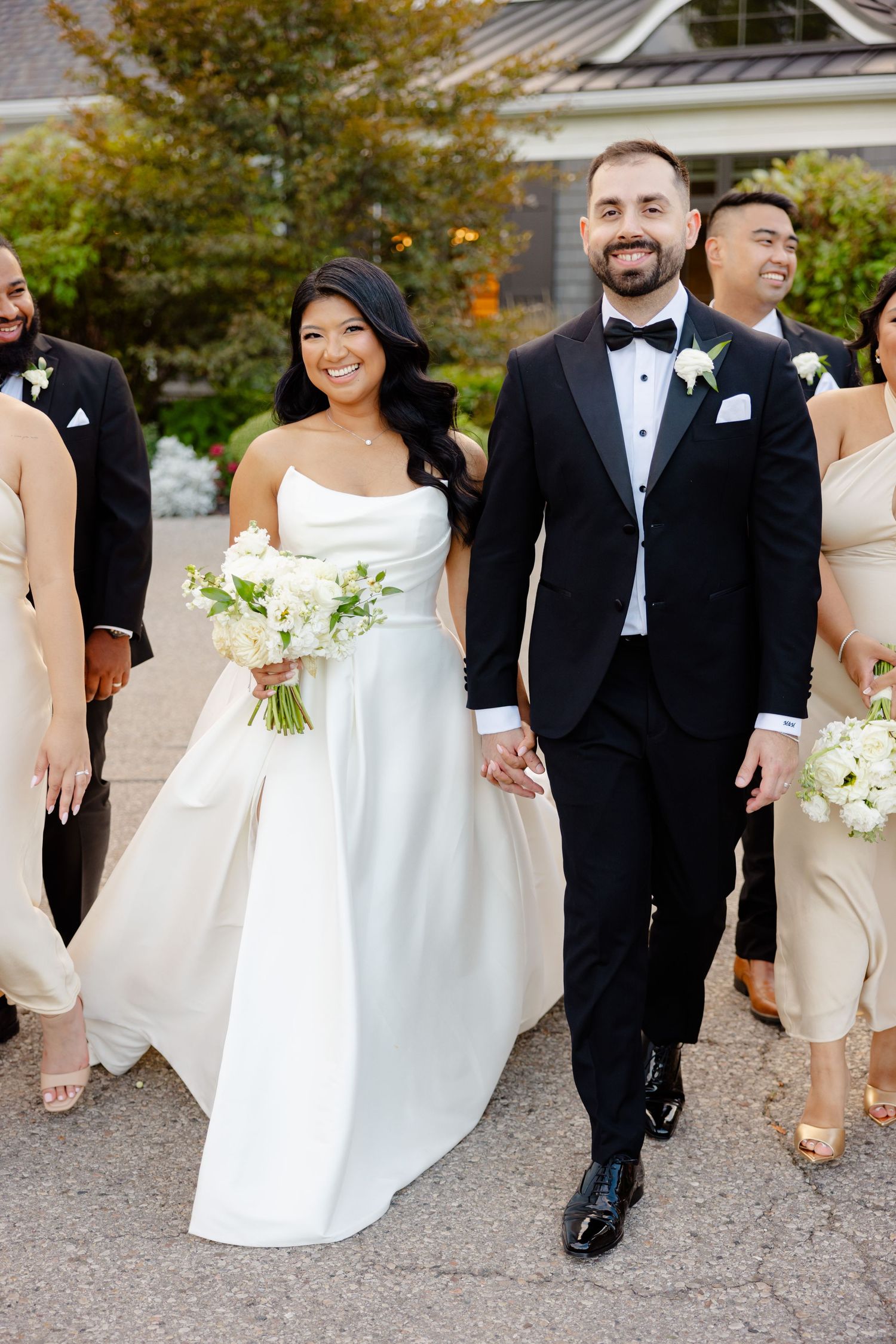 Wedding couple and party members walk together outside gray building with autumn foliage.