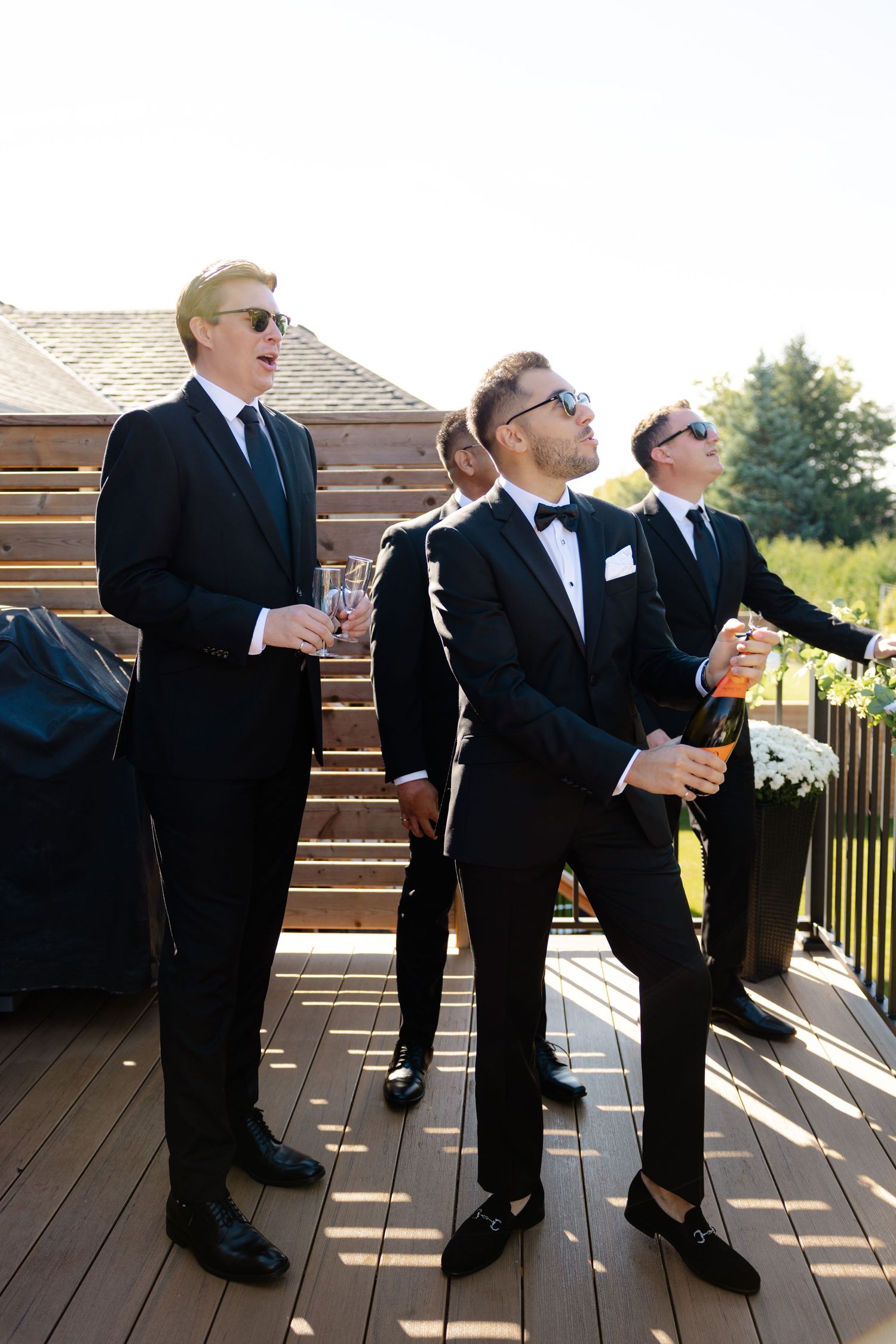 Groomsmen in formal attire holding drinks while standing on outdoor deck.