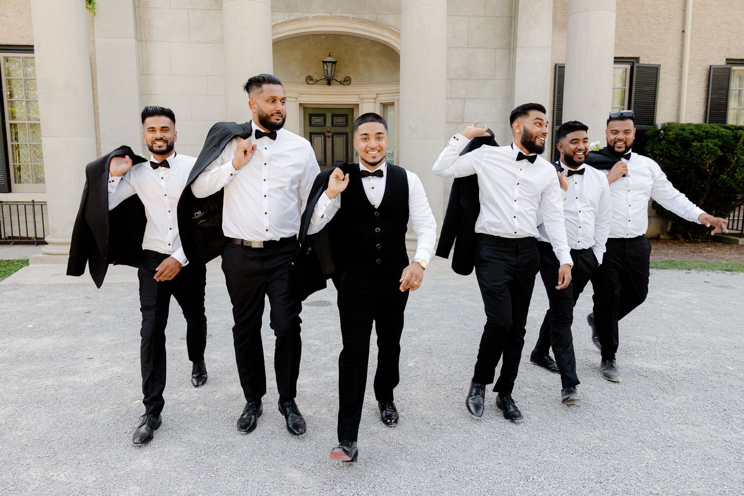 Groomsmen in black and white formal attire jumping and posing playfully outside a stately building.