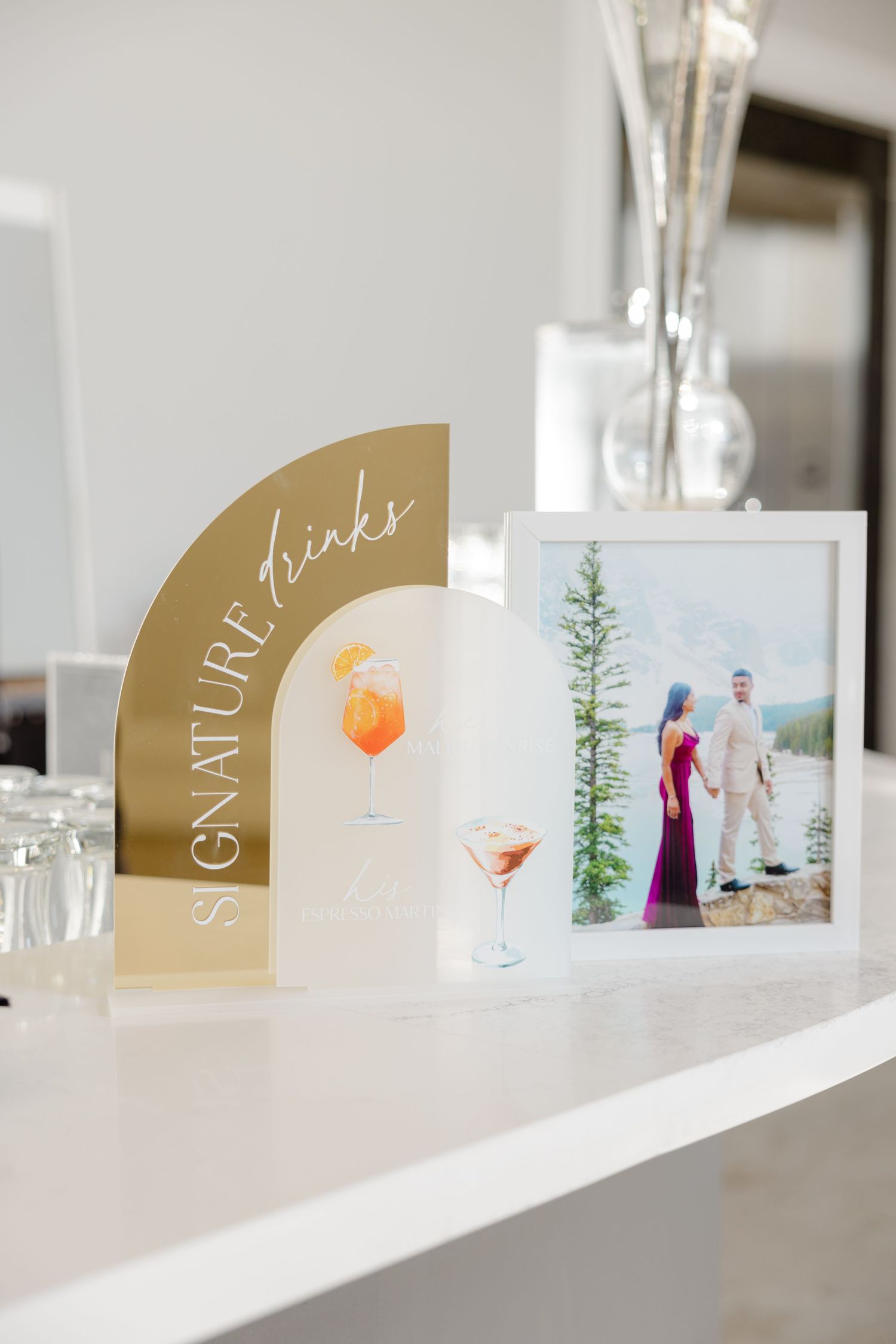 Gold signature drinks menu display with cocktail illustrations and portrait on white table.