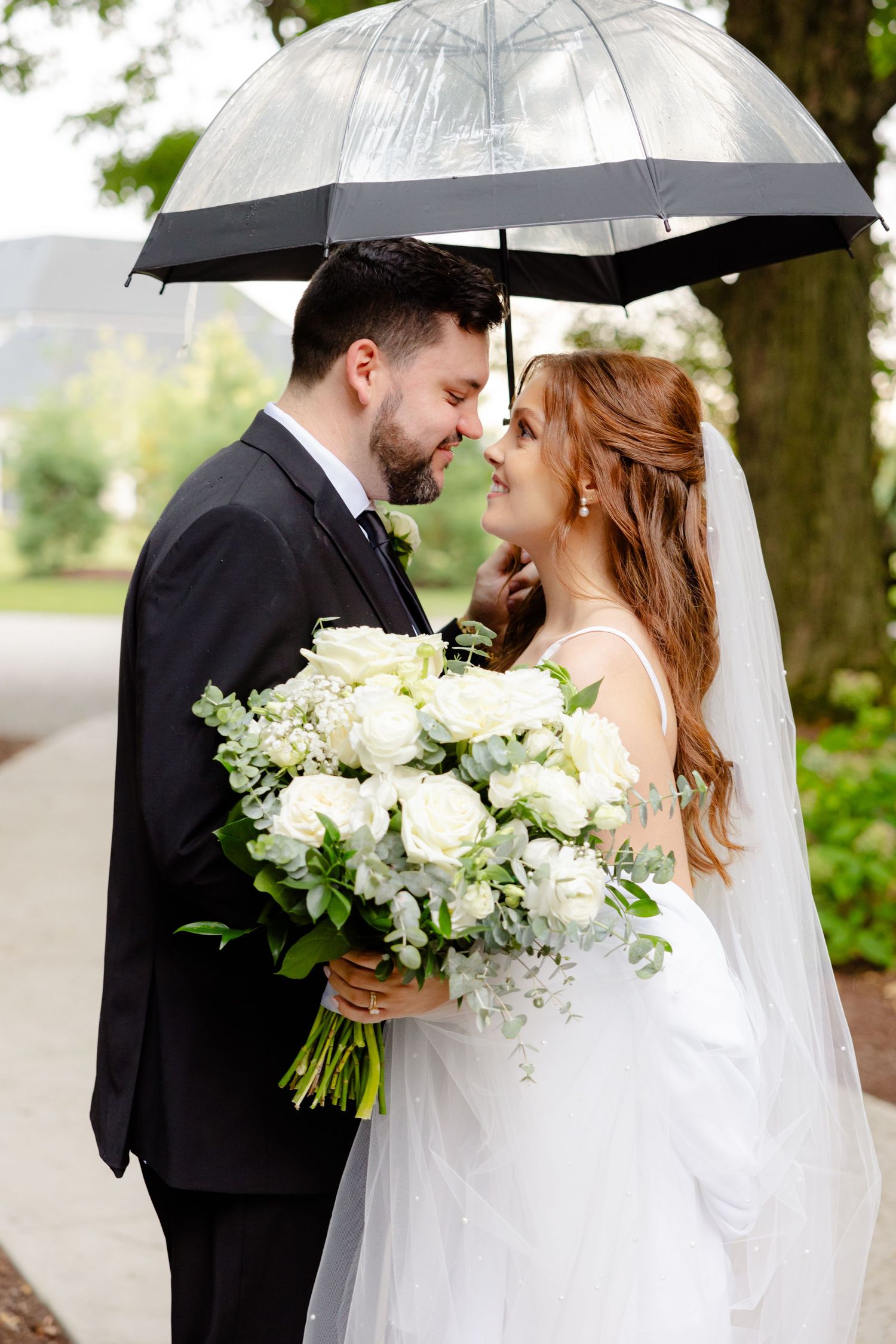 Newlyweds share a romantic moment beneath a transparent umbrella while holding a lush white bouquet.
