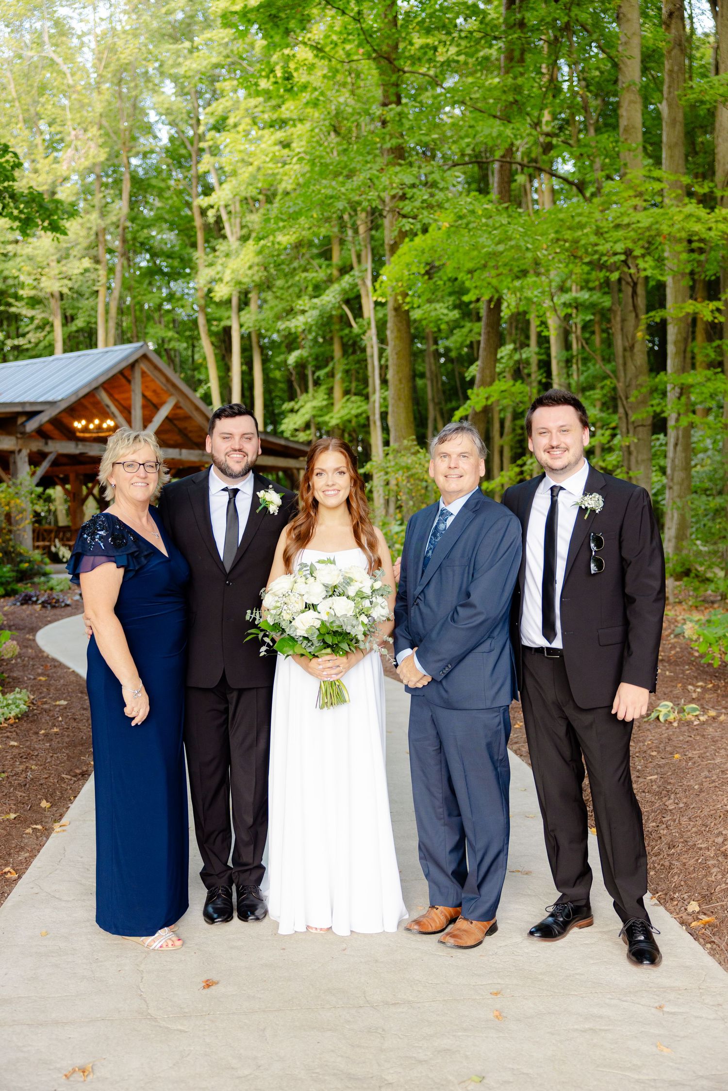 Formal wedding party group photo in front of a wooden pavilion surrounded by lush green forest.