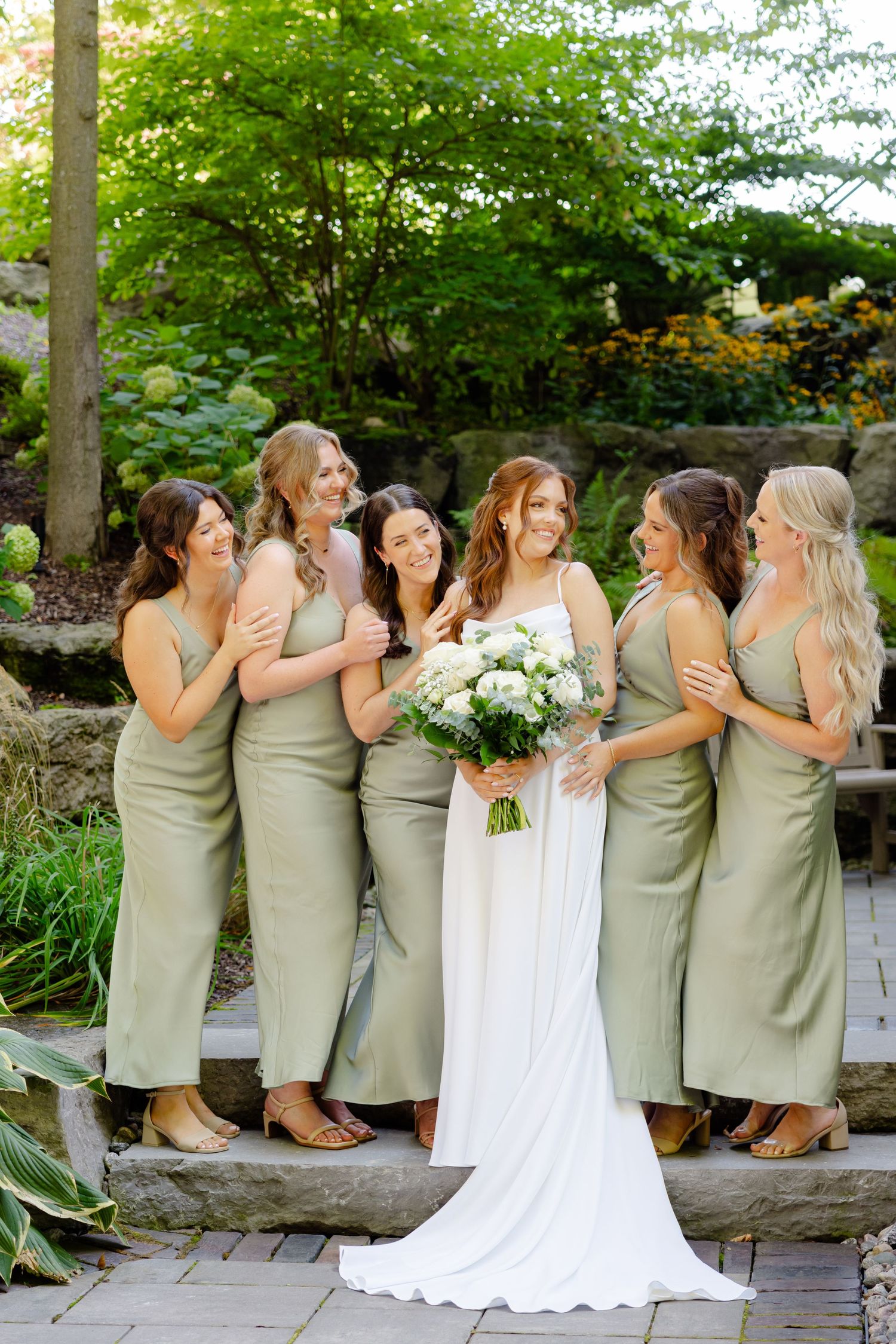 Wedding party shares joyful moment on stone steps with bridesmaids in sage and bride in flowing white gown.