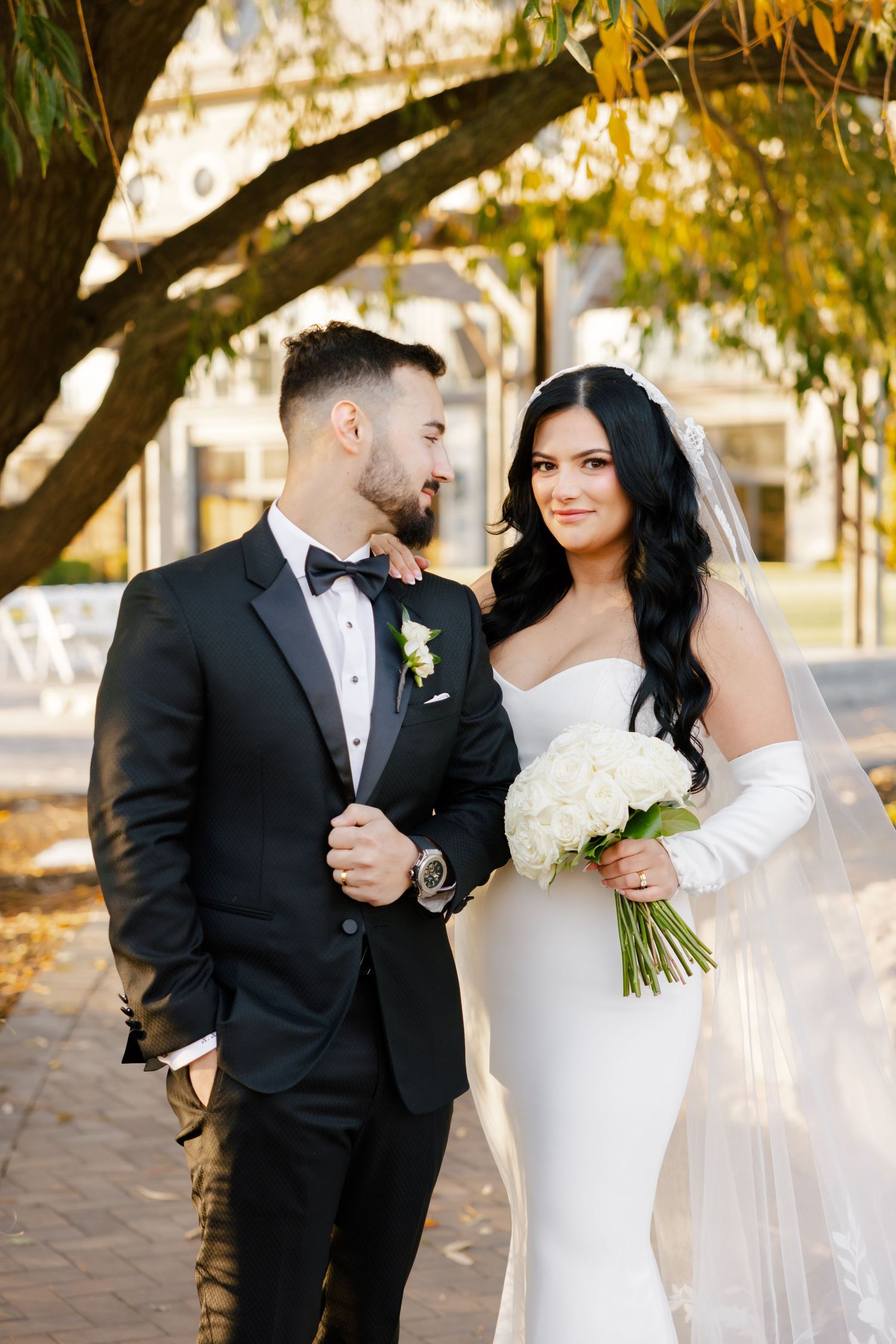 Couple in formal wedding attire stands together under golden autumn tree branches.