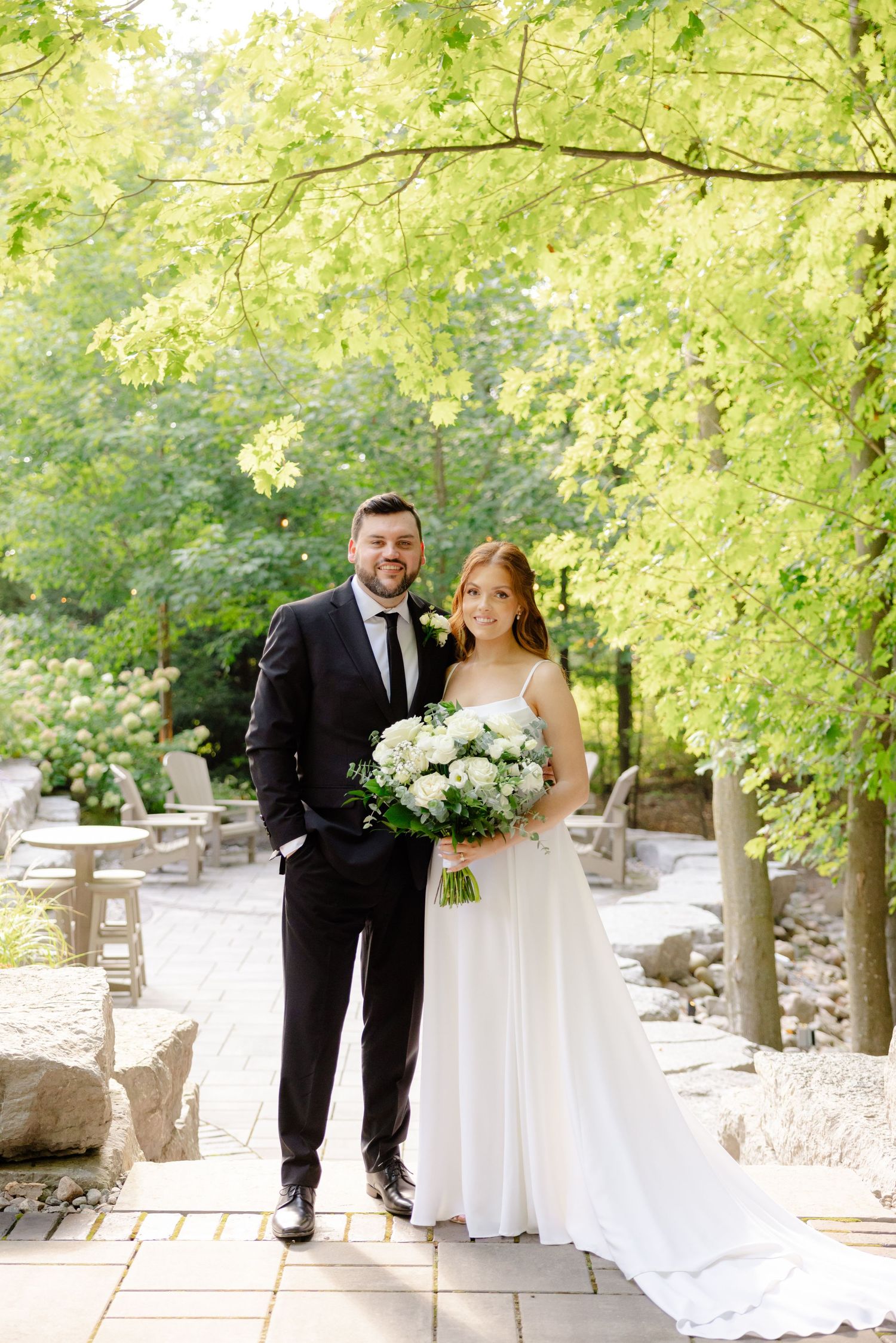Elegant wedding portrait on stone pathway under green leafy canopy.